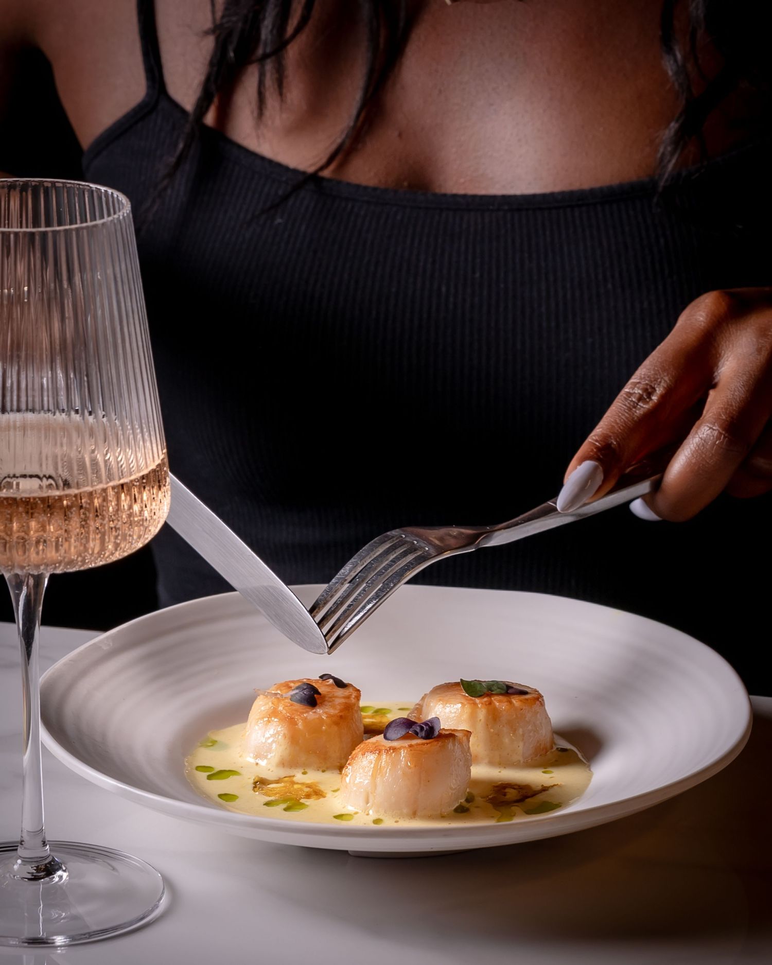 Woman in black top dining on seared scallops in butter sauce, with a glass of rosé wine at an elegant restaurant.
