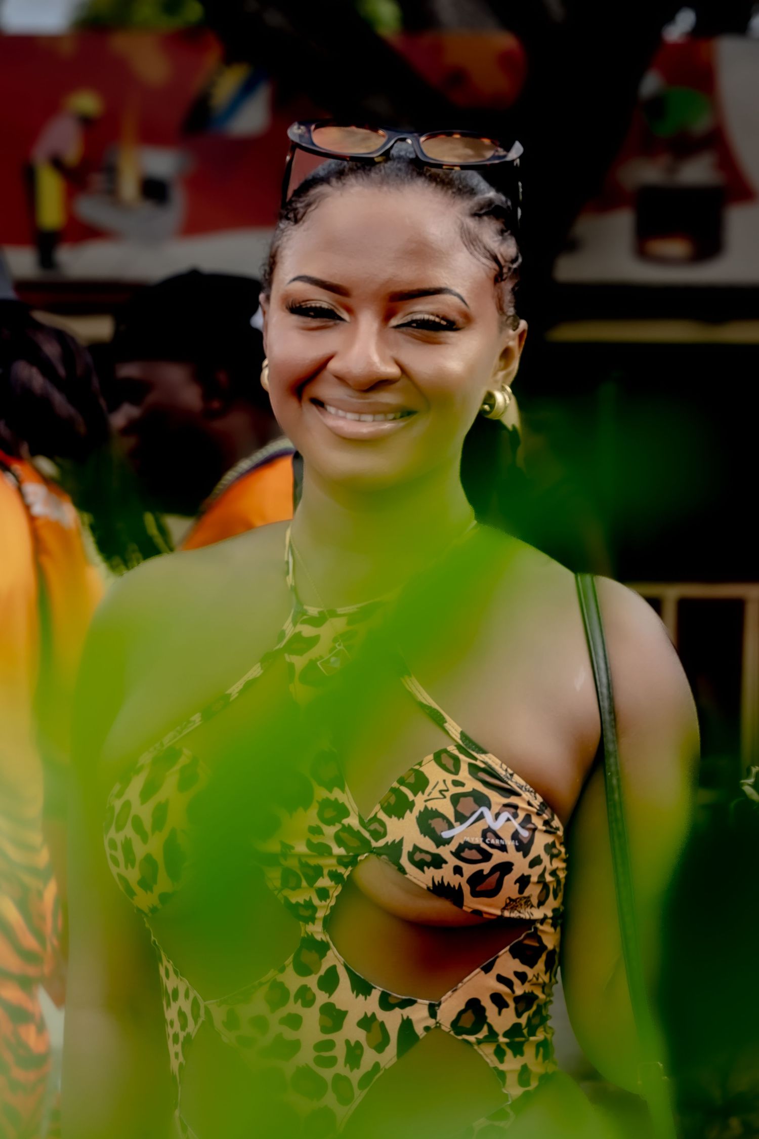 A smiling woman in a leopard print bikini top with sunglasses on her head at a colorful outdoor carnival event.