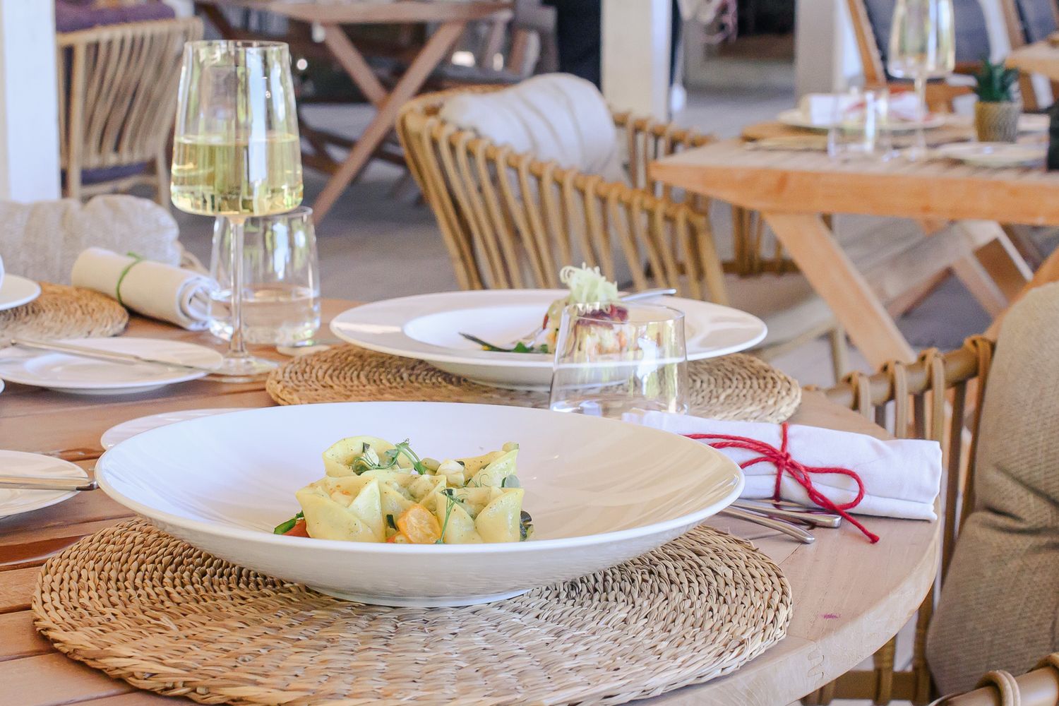 Elegant table settings at a restaurant with woven placemats, white plates, and wine glasses in natural lighting.