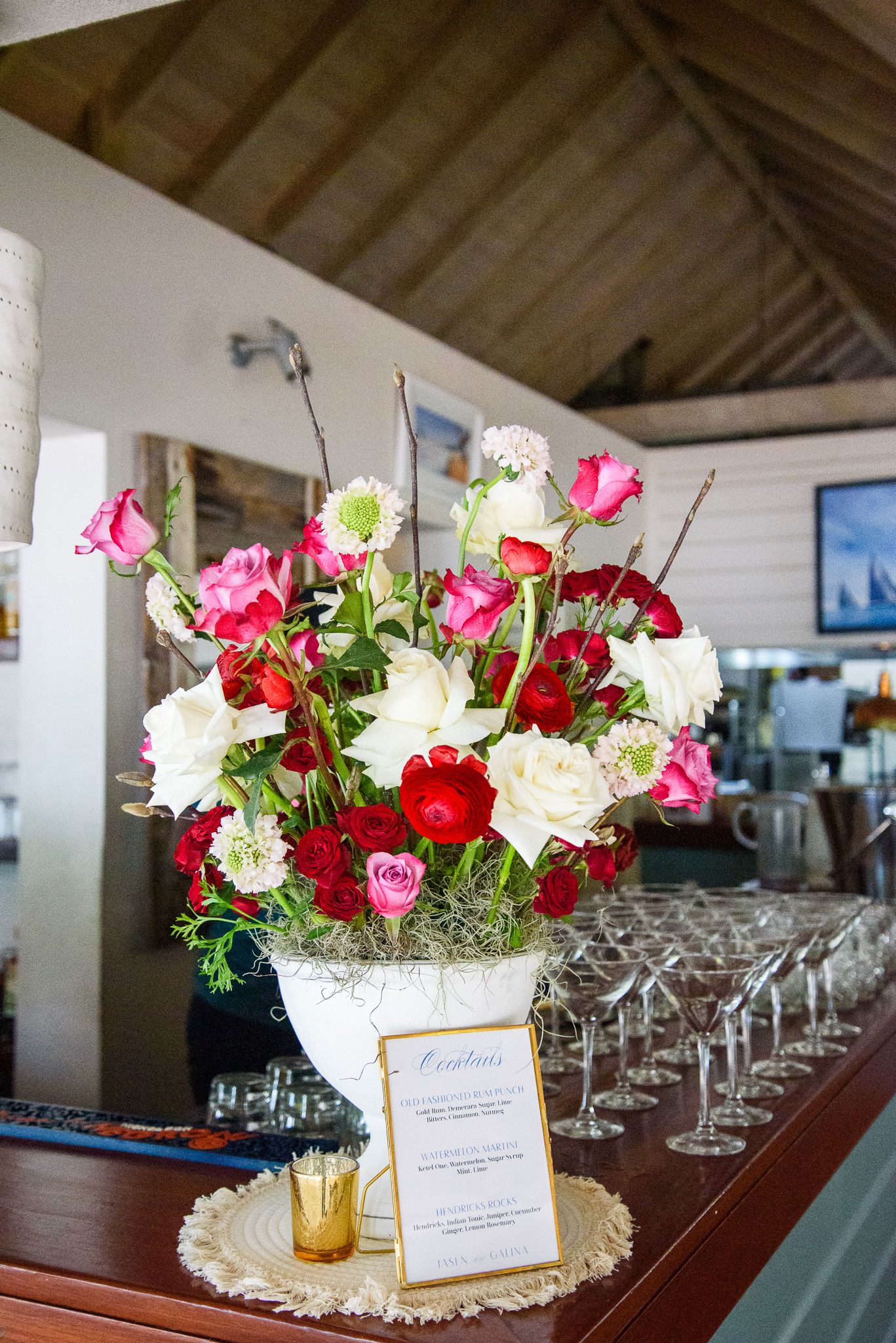 Wedding reception table centerpieces featuring red and pink roses in gold vases with place settings.