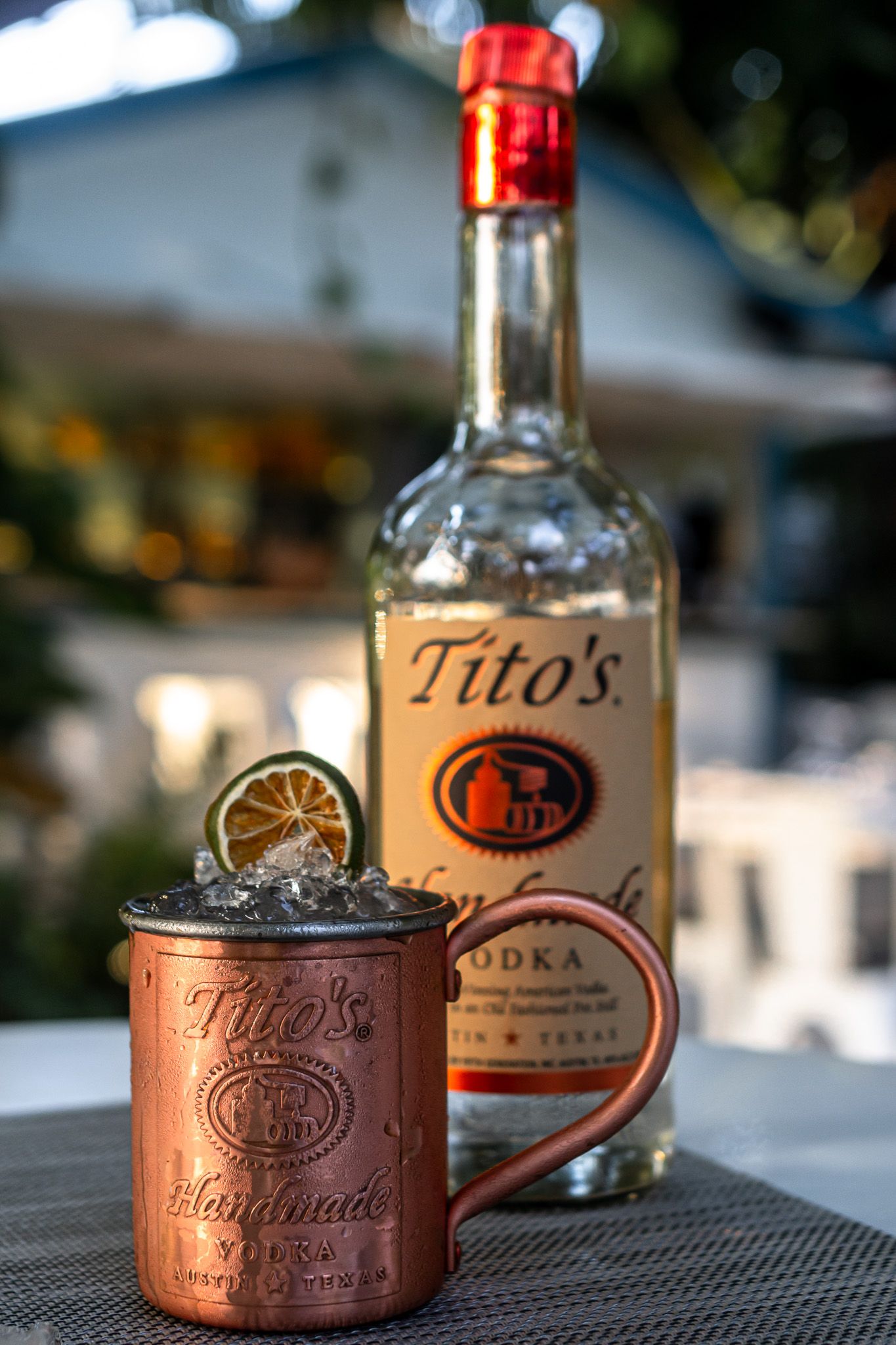 A copper Moscow Mule mug garnished with lime sits next to a bottle of Tito's Handmade Vodka on an outdoor table.