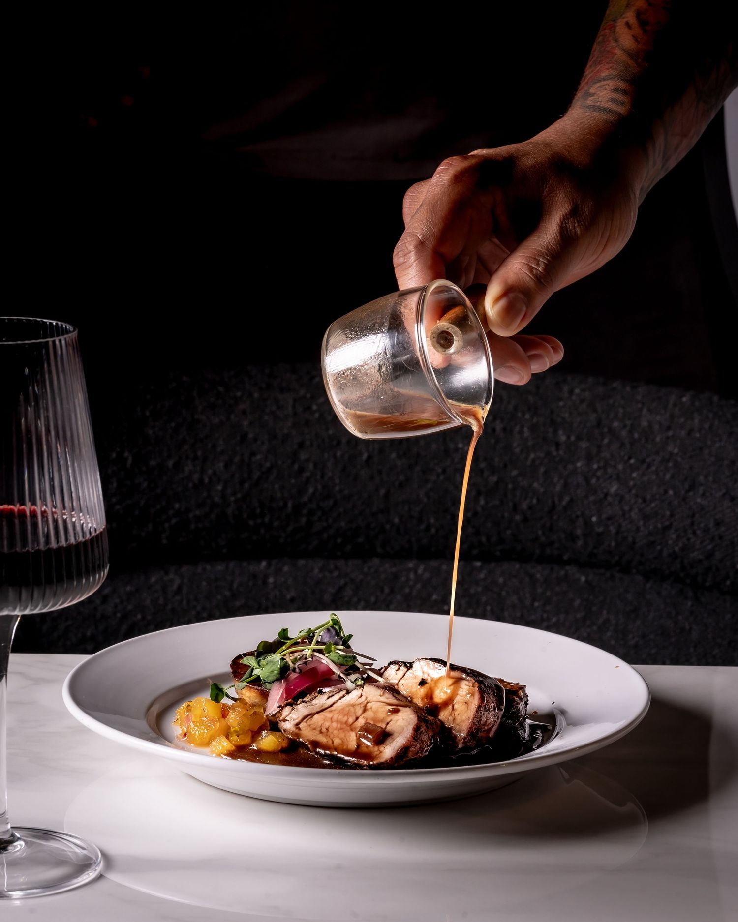 Chef pouring rich sauce over an elegant plated meat dish with garnish, served with red wine at a fine dining restaurant.