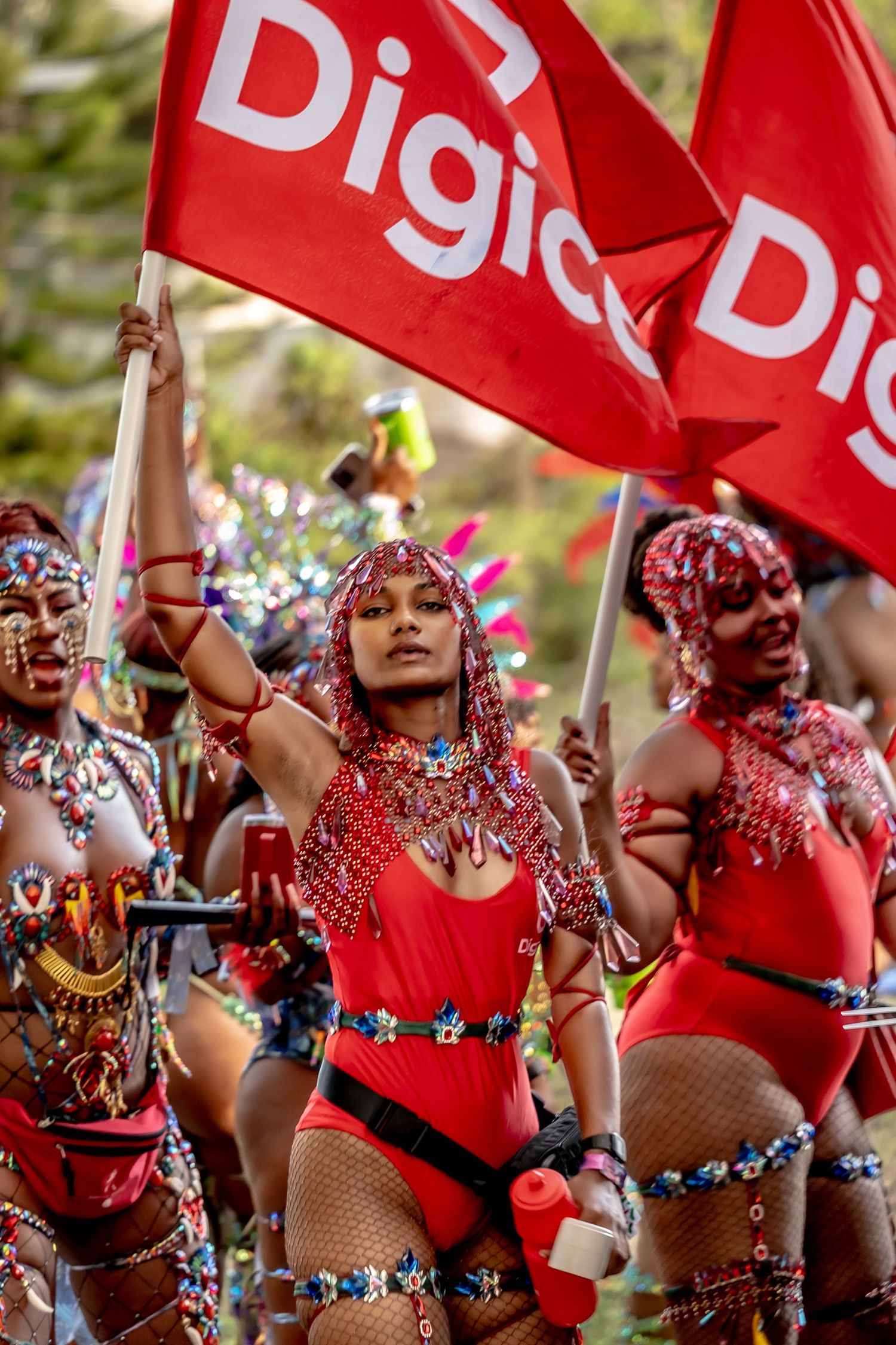 Women in red beaded costumes carry Digicel flags during a vibrant Caribbean carnival parade celebration.