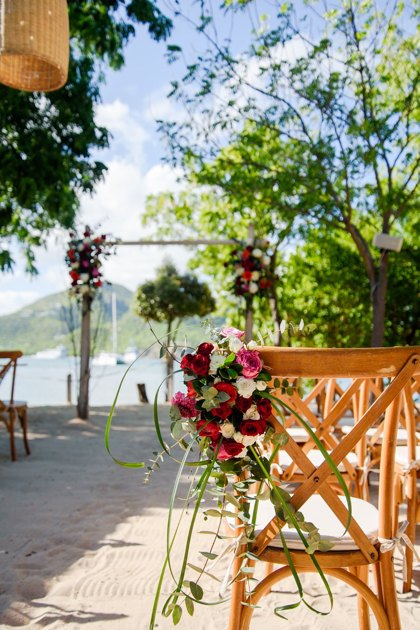 Wooden chairs decorated with burgundy and pink floral arrangements line up on a sandy beach for an outdoor wedding ceremony.
