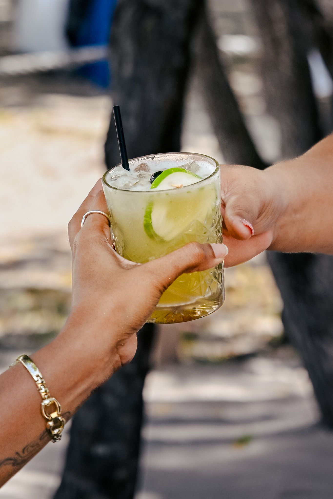 Two hands clink margarita glasses garnished with lime slices in a celebratory outdoor toast.