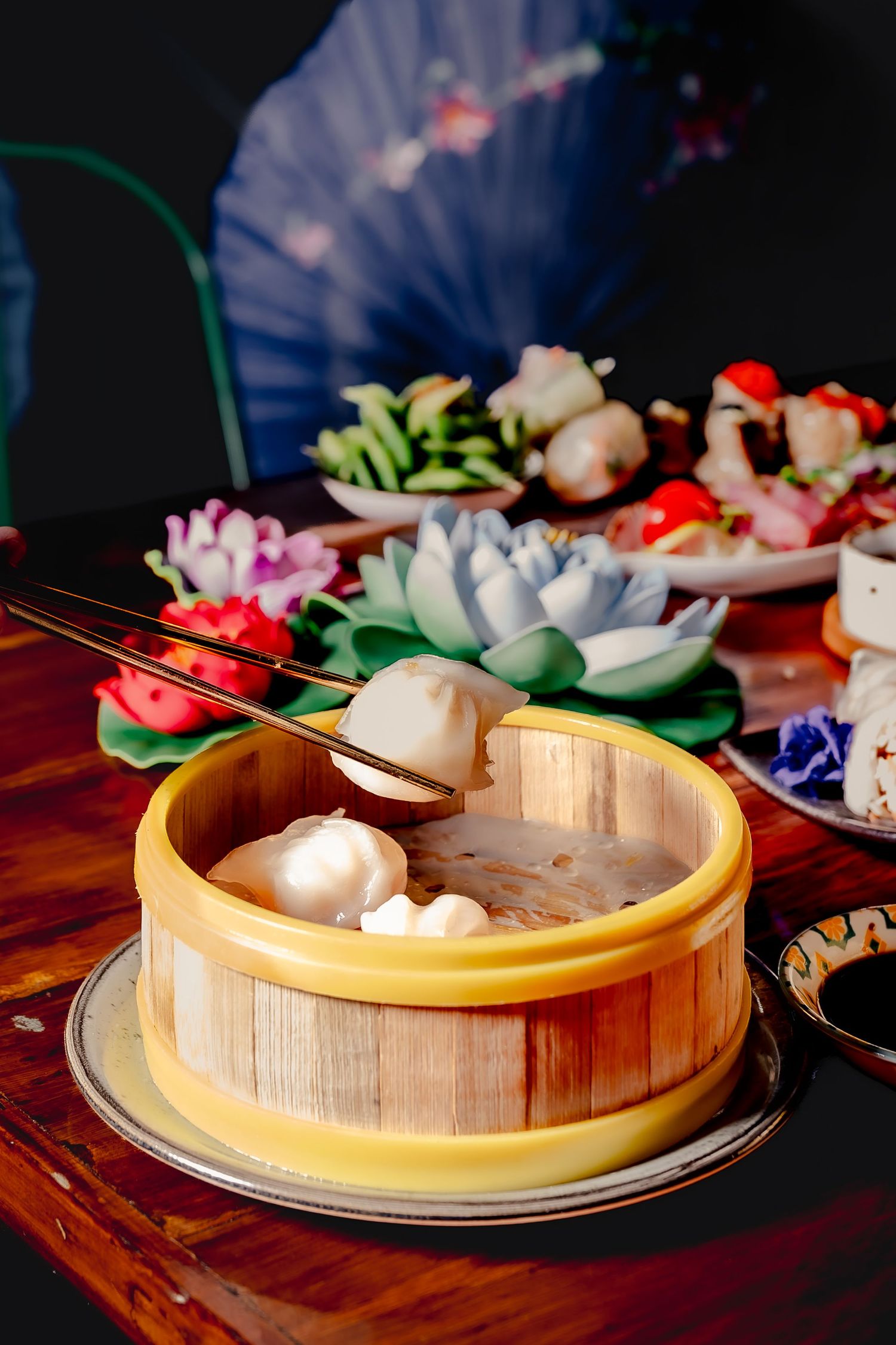 Bamboo steamer basket with dim sum dumplings on a wooden table, surrounded by colorful decorative flowers and dishes.