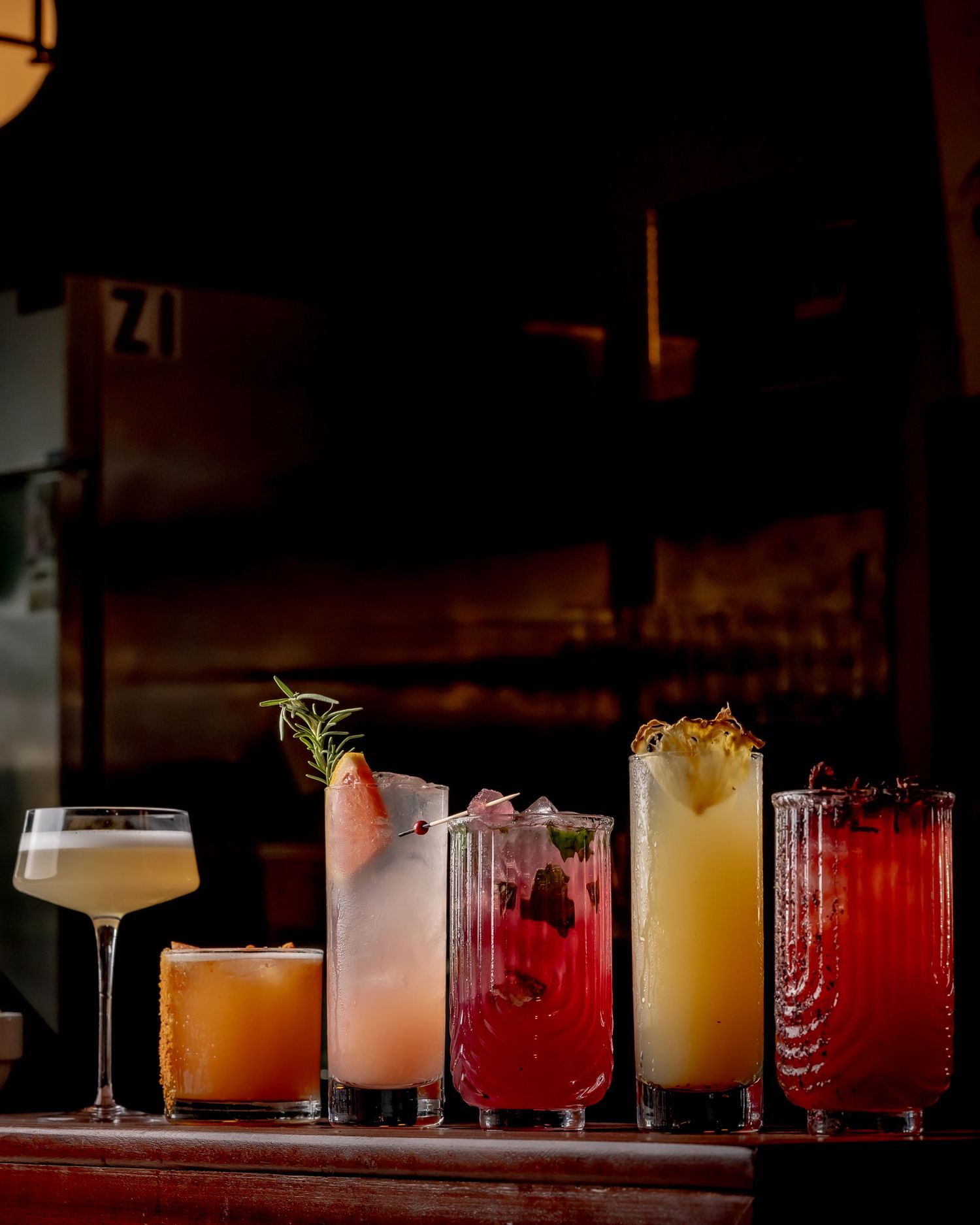 Five colorful cocktails lined up on a dimly lit bar, featuring garnishes like rosemary, fruit, and pineapple.