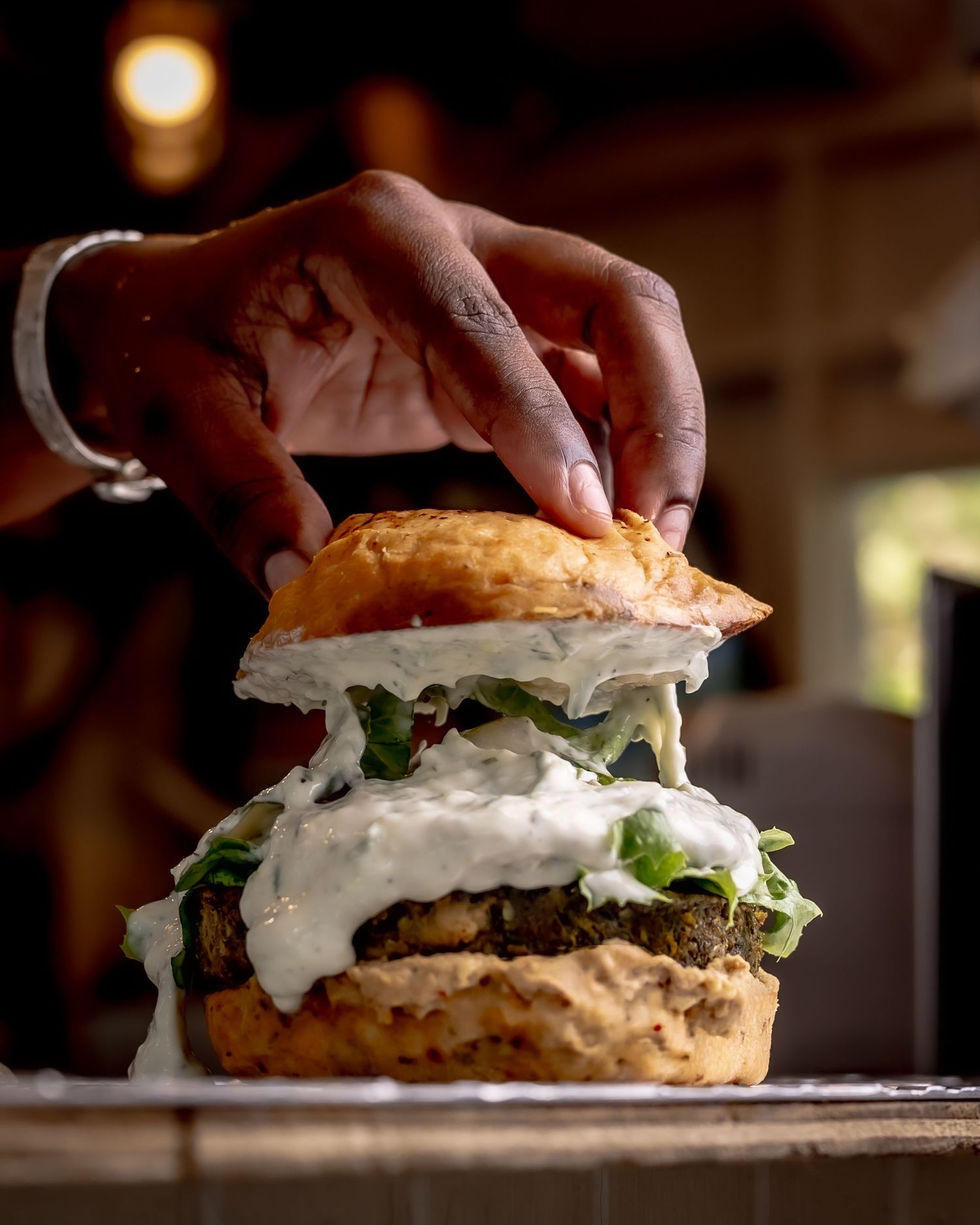 A hand holds a towering gourmet burger topped with white sauce and fresh lettuce on a rustic wooden surface.