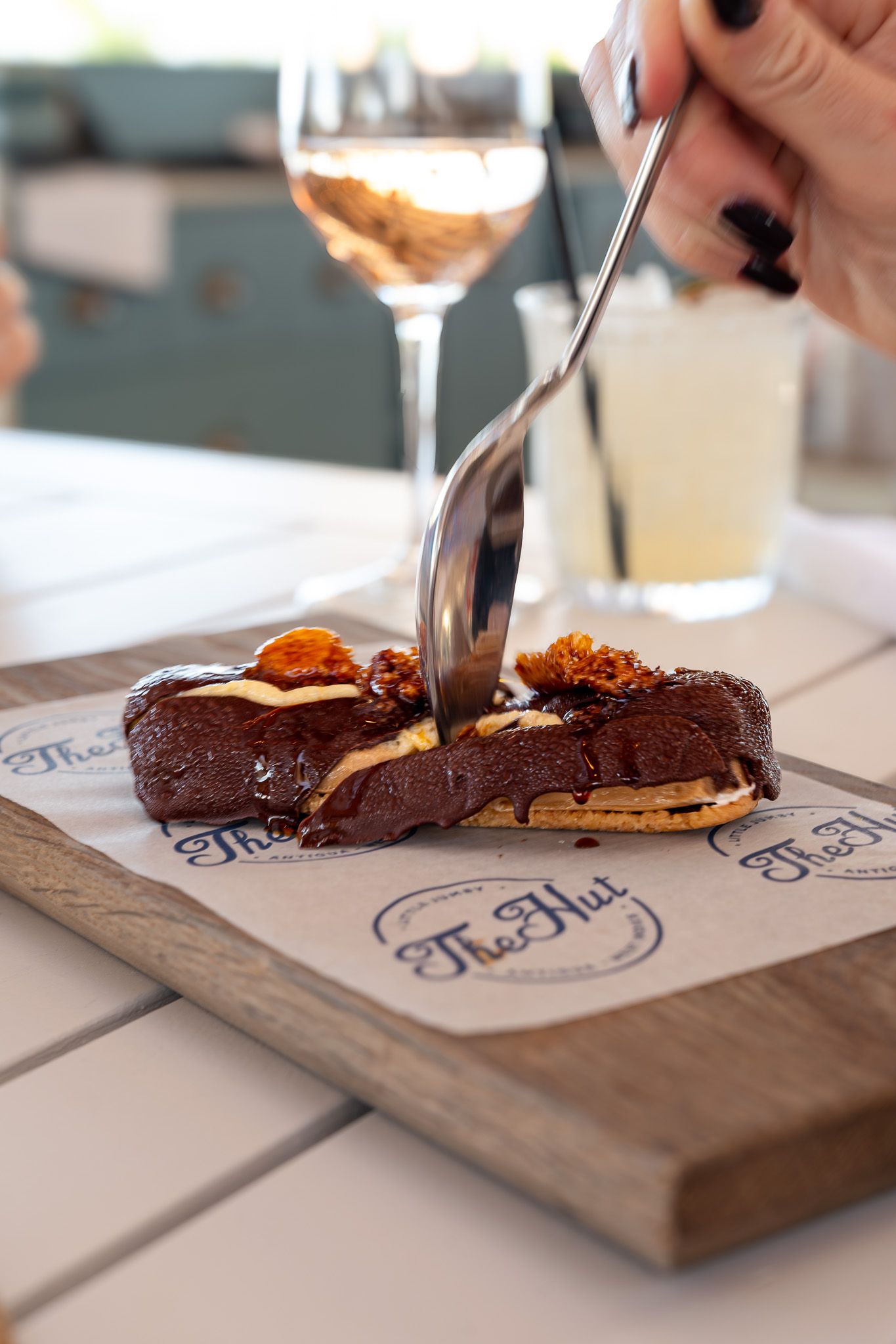 Rich chocolate dessert with caramel and nuts being cut on a wooden serving board with wine glass in background.