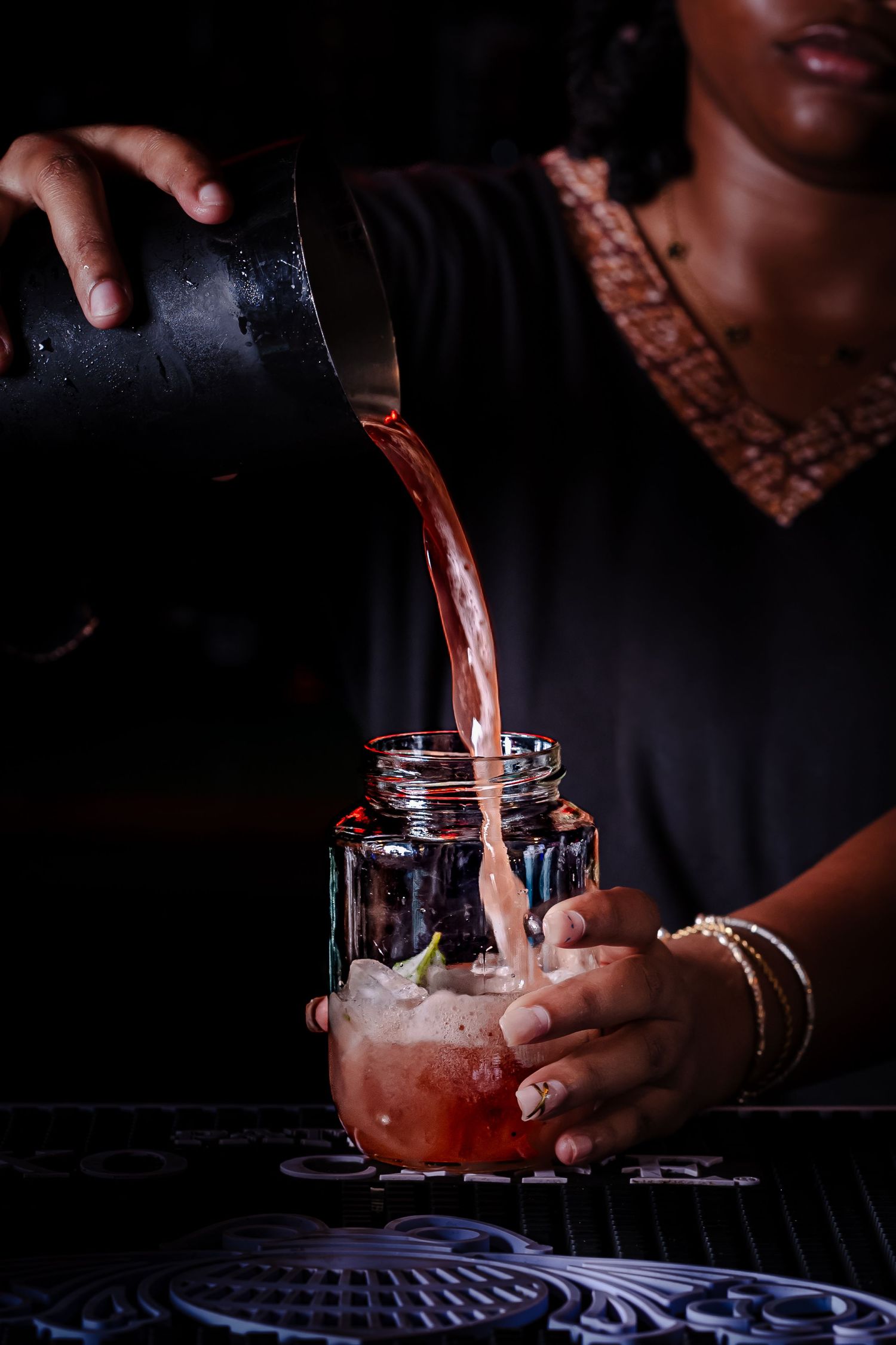Bartender pouring pink cocktail into mason jar glass with ice and fresh herbs at a bar.