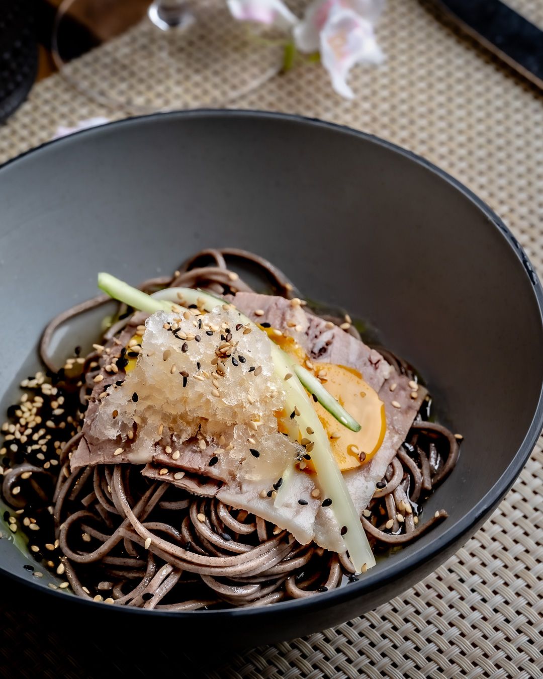 A black bowl of soba noodles topped with sliced meat, cucumber, egg, sesame seeds, and a savory sauce.