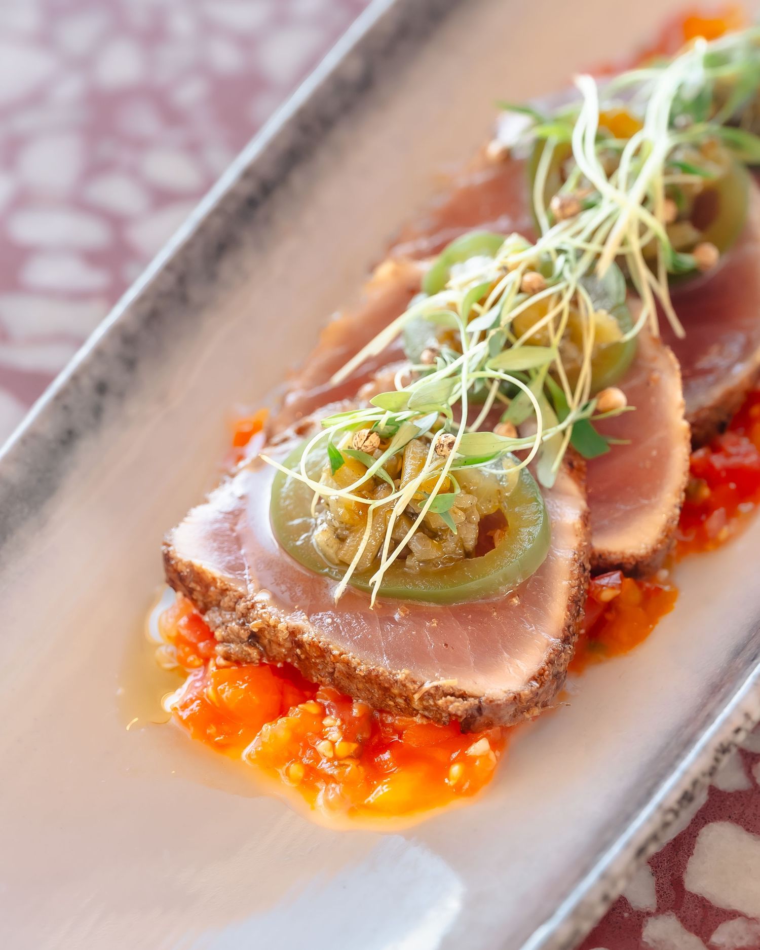 Seared ahi tuna slices on tomato sauce, topped with jalapeno rounds and microgreens on a white rectangular plate.
