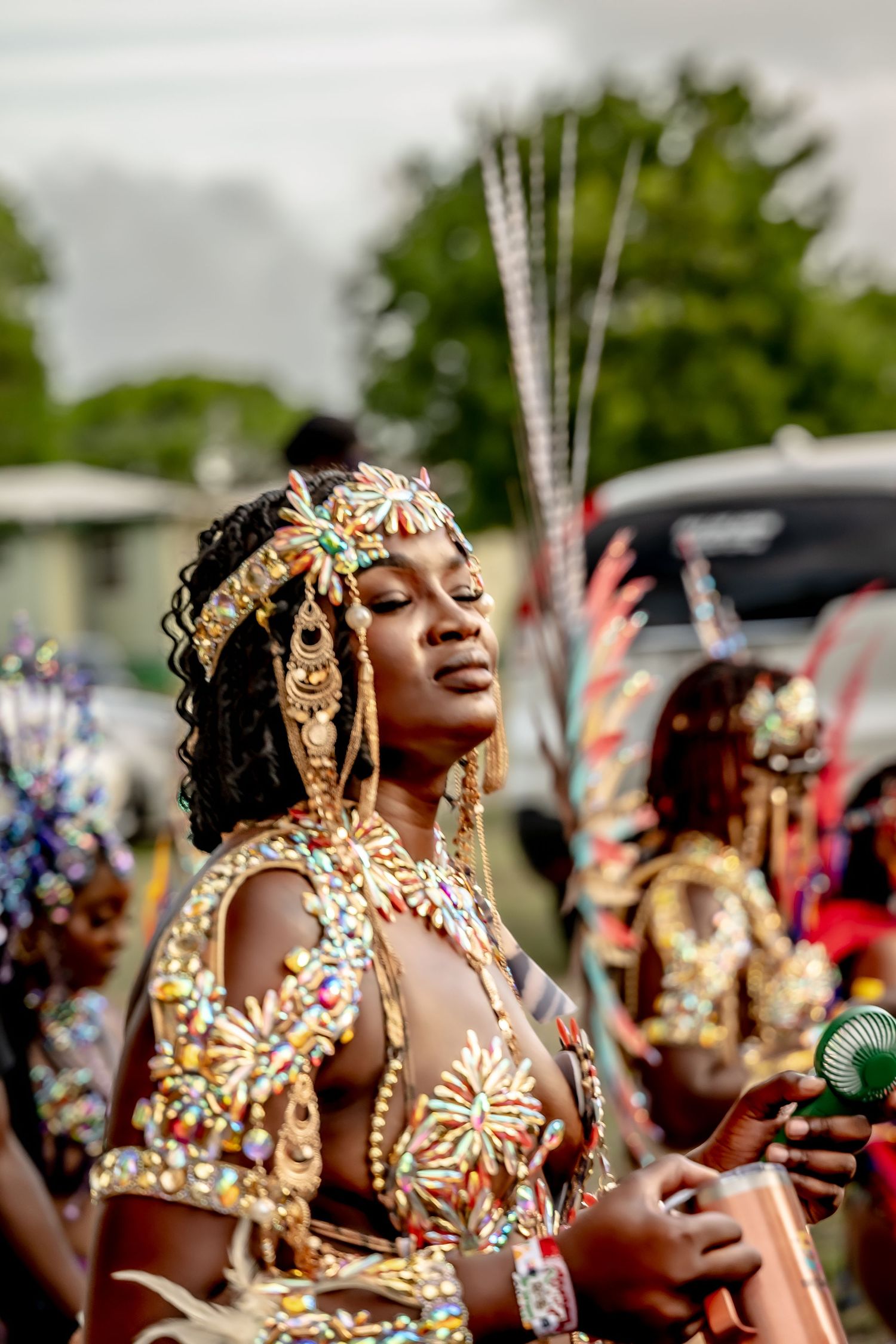 A woman in ornate gold jeweled carnival costume with feathered headdress poses gracefully at a vibrant Caribbean festival.