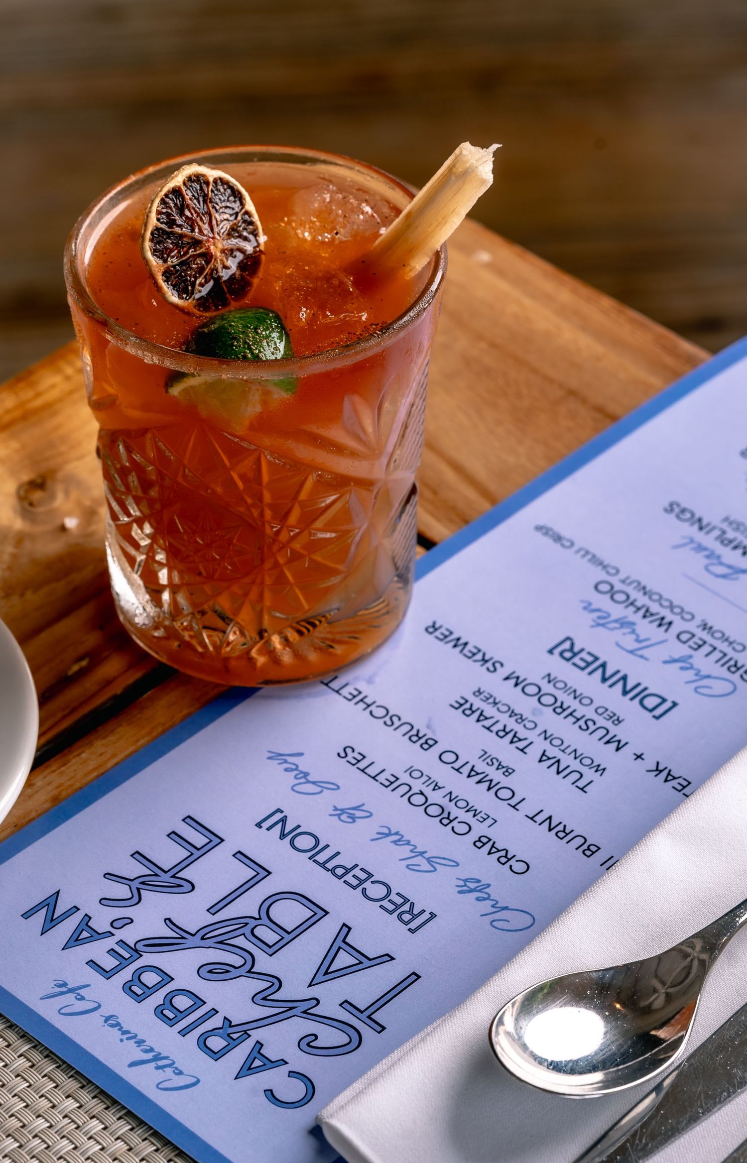 A cocktail with ice, dried citrus, and a cinnamon stick sits on a wooden board beside a Caribbean Table dinner menu.