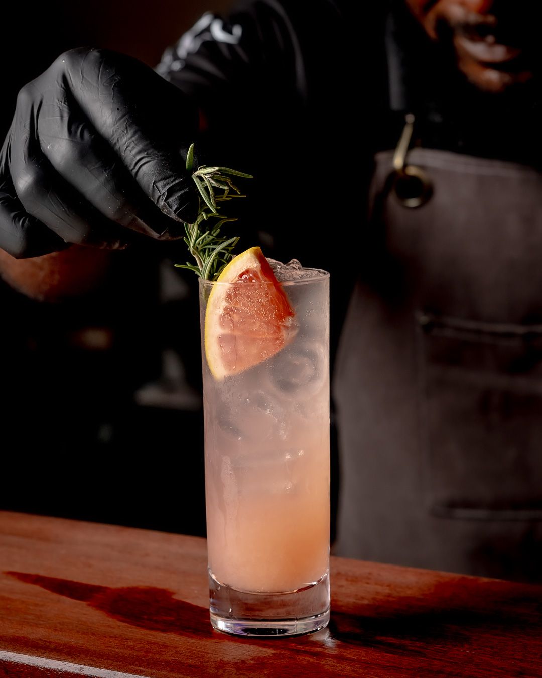 Bartender in black gloves garnishing a pink grapefruit cocktail with rosemary in a tall glass on a wooden bar.