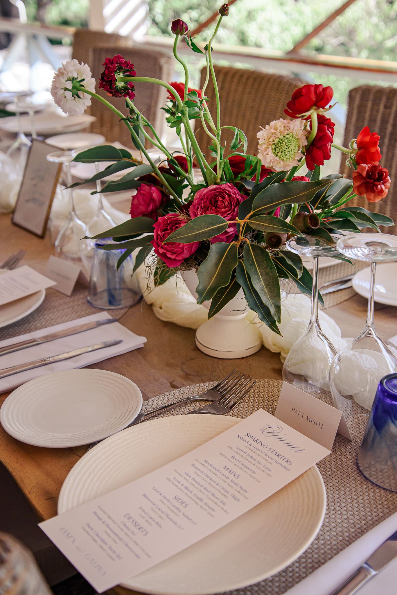 Table settings with white plates, menus and floral arrangements featuring red roses and greenery.