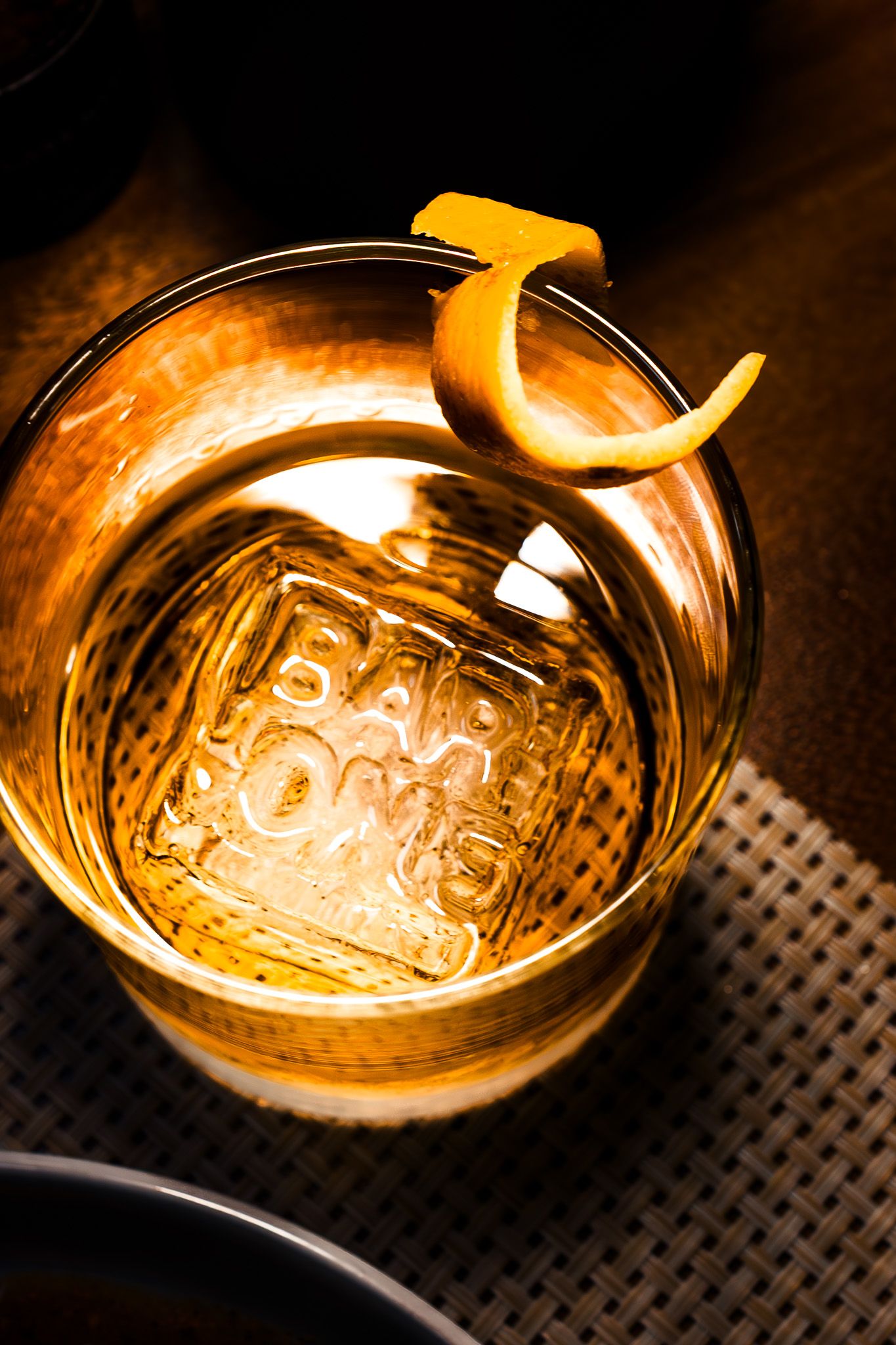 Cocktail glass with large ice cube and orange peel garnish glowing with warm amber lighting on textured surface.