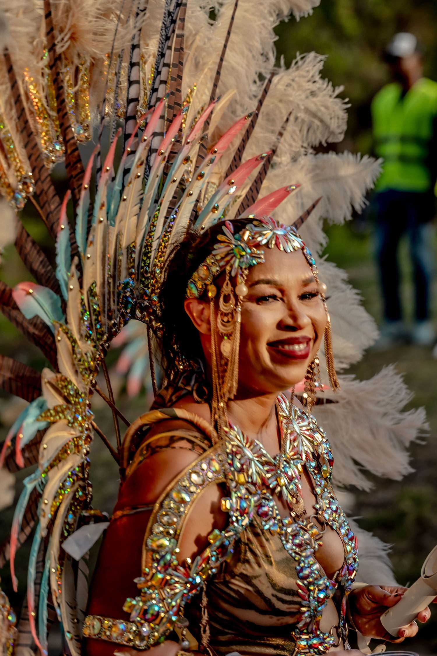 Smiling woman in ornate carnival costume with feathered headdress and jeweled outfit at a festive parade.