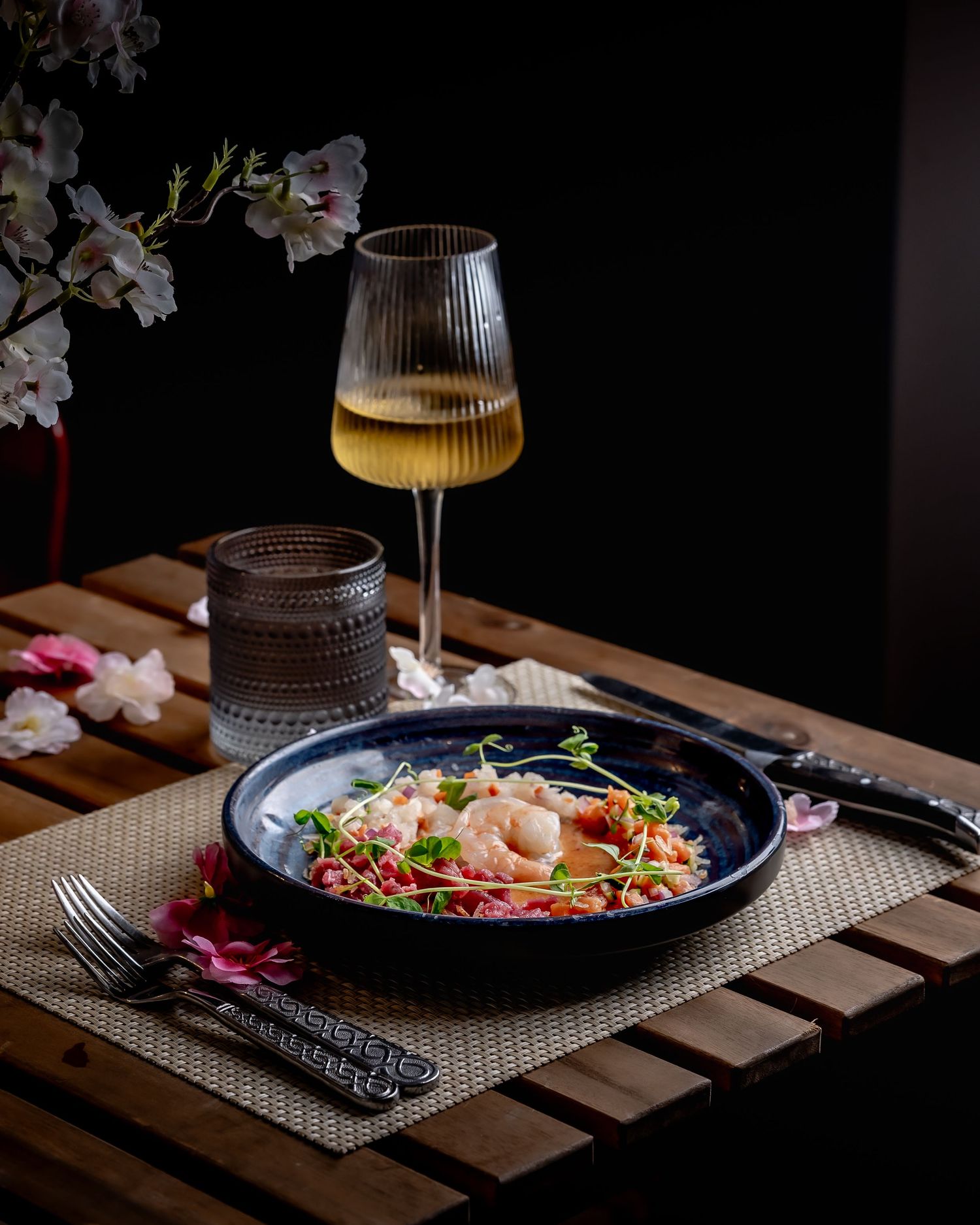Elegant shrimp pasta dish in dark bowl with white wine glass and flowers on wooden table.
