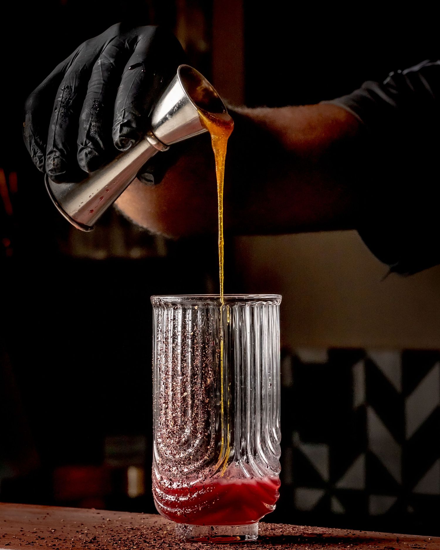 Bartender in black gloves pouring amber liquid from a jigger into a ribbed glass with red cocktail base.