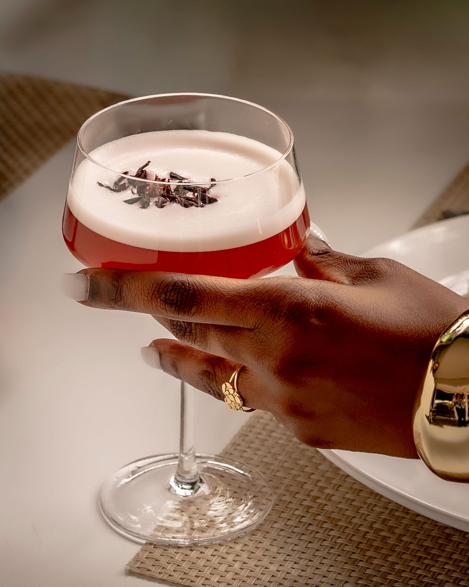 A hand with gold jewelry holds an elegant coupe glass with a pink cocktail topped with dried flowers.