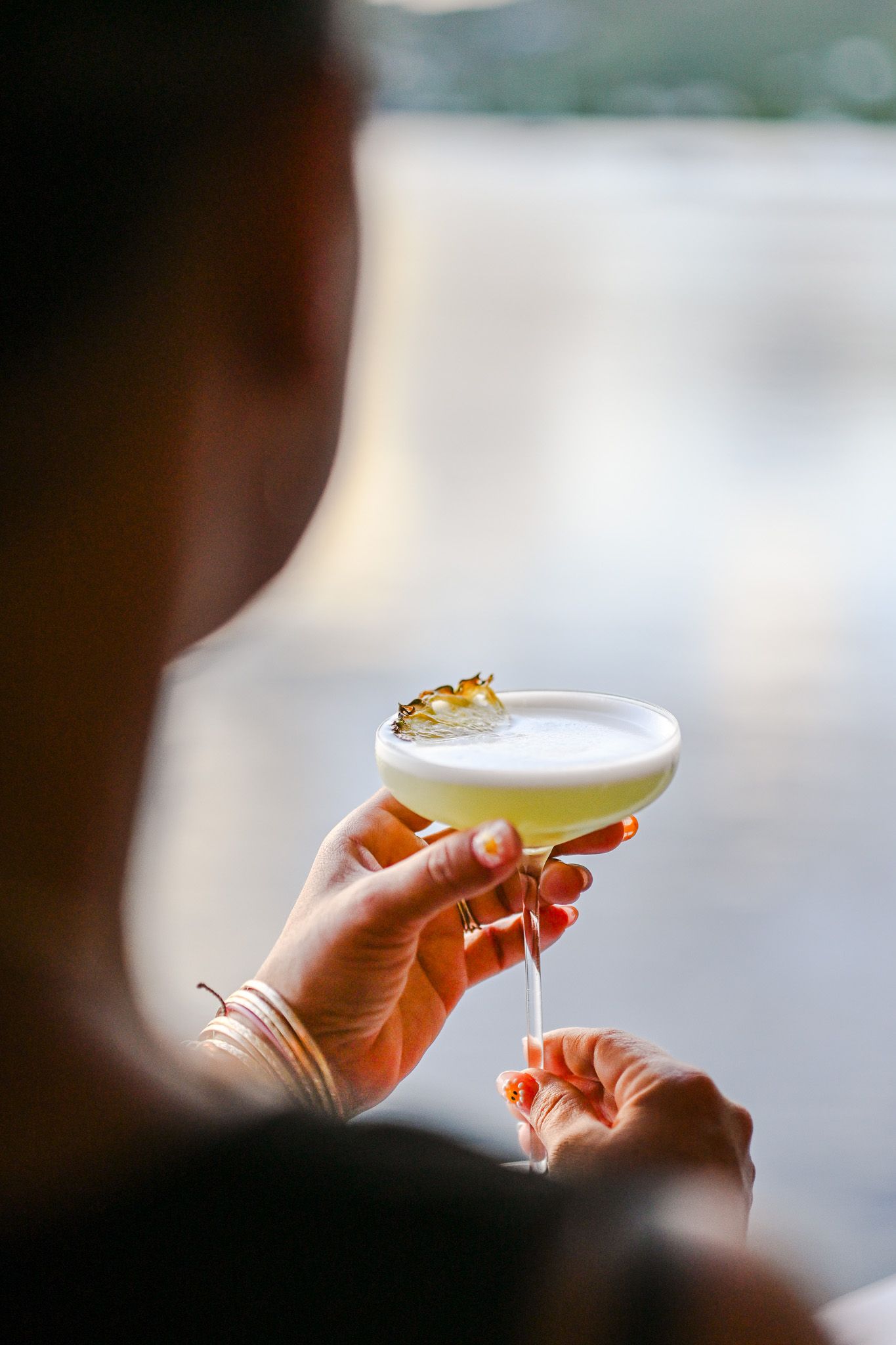 Person holding a frothy pale yellow cocktail in a coupe glass garnished with a pineapple slice.