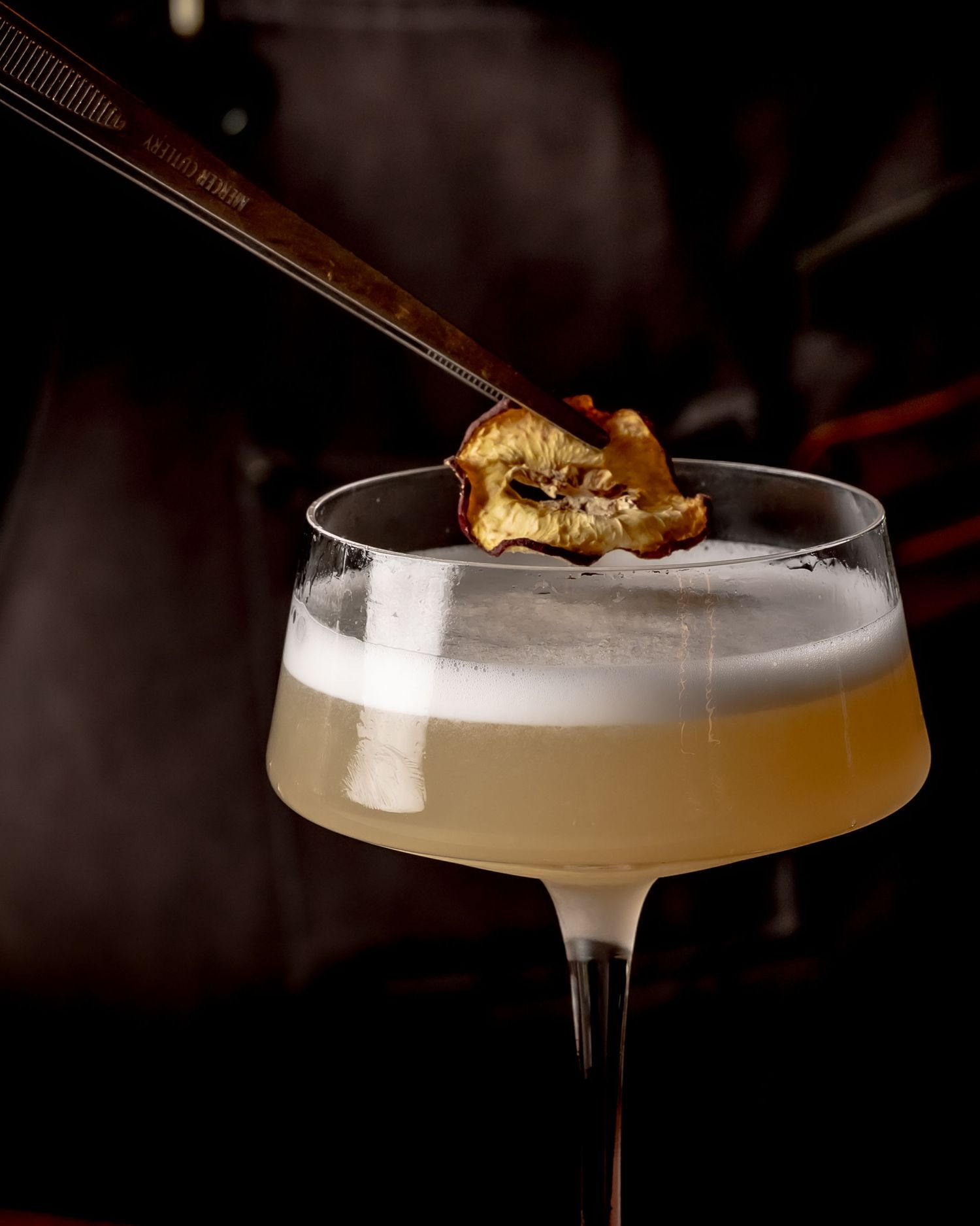 Elegant cocktail in a coupe glass with frothy top, garnished with a dried fruit piece held by tweezers.
