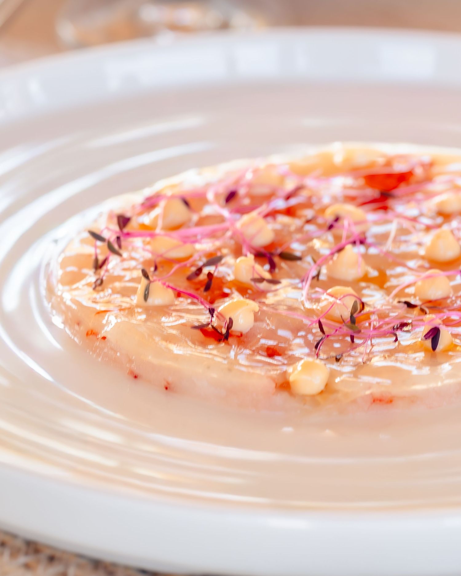 Elegant carpaccio dish with floral garnish, toasted nuts, and delicate broth served on a white ceramic plate.