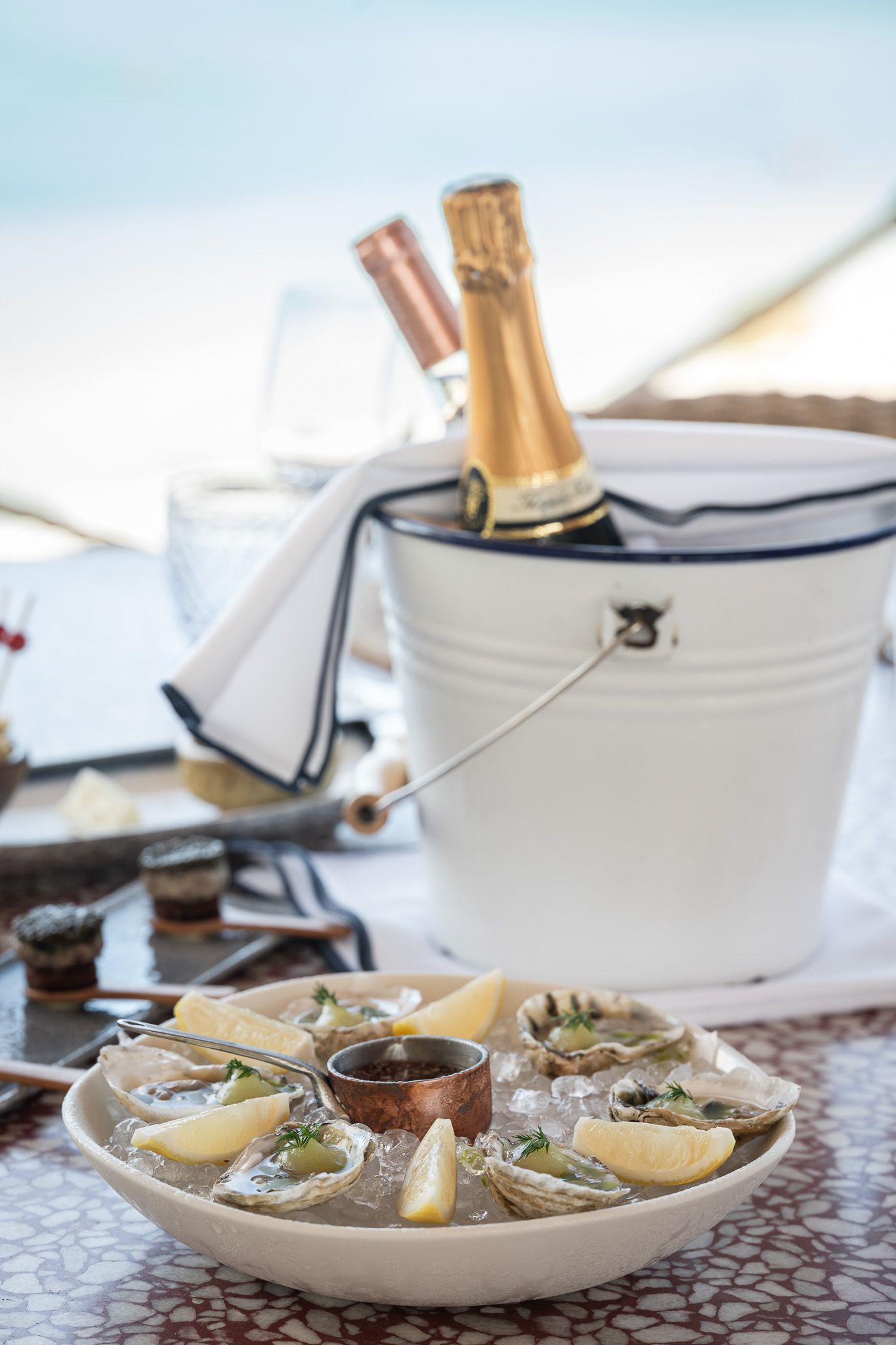 Fresh oysters on ice with lemon wedges served in white ceramic plates alongside chilled champagne bottles in ice buckets.