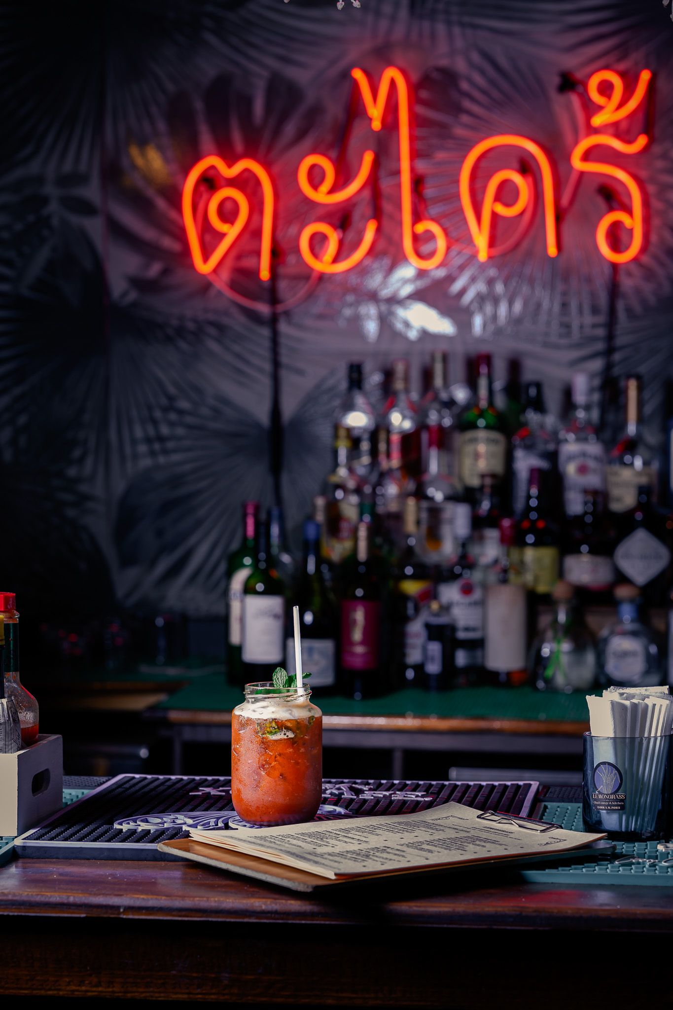 Thai bar with neon sign, cocktail in mason jar on wooden counter, liquor bottles displayed on shelves behind.