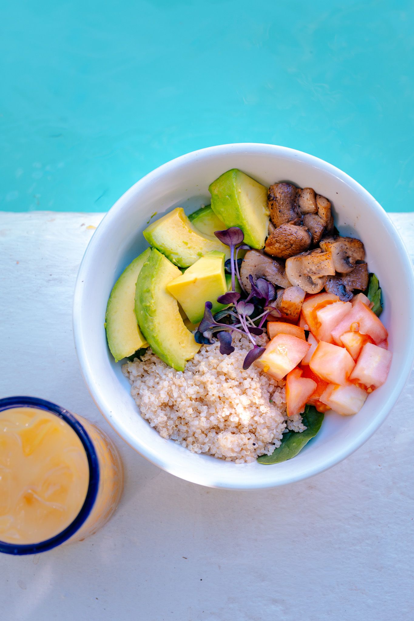 A vibrant Buddha bowl with avocado, quinoa, mushrooms, tomatoes, and microgreens beside a mango smoothie by a pool.