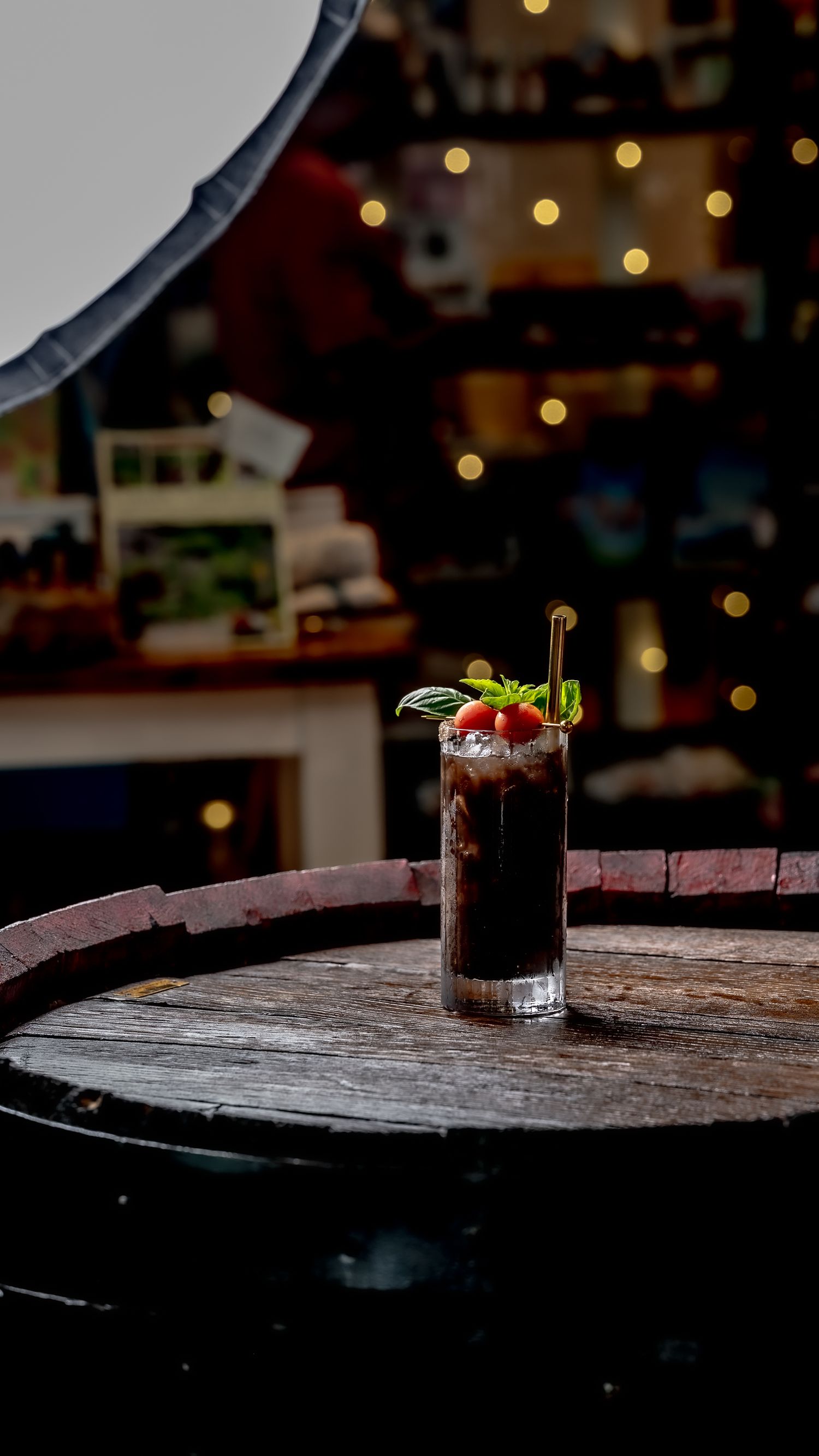 Dark cocktail garnished with mint and berries in a tall glass on a rustic wooden barrel in a warmly lit bar.