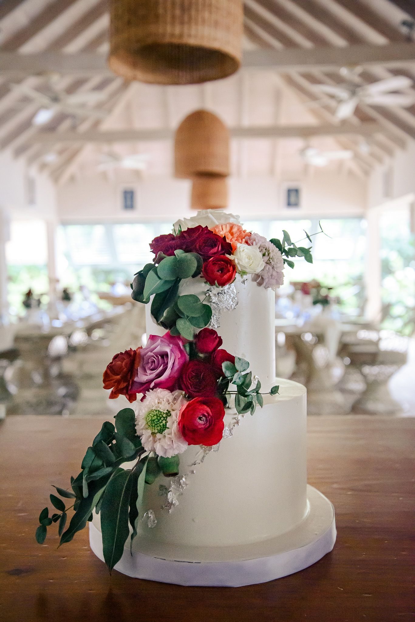 White tiered wedding cake decorated with cascading red and pink roses and greenery on wooden table.