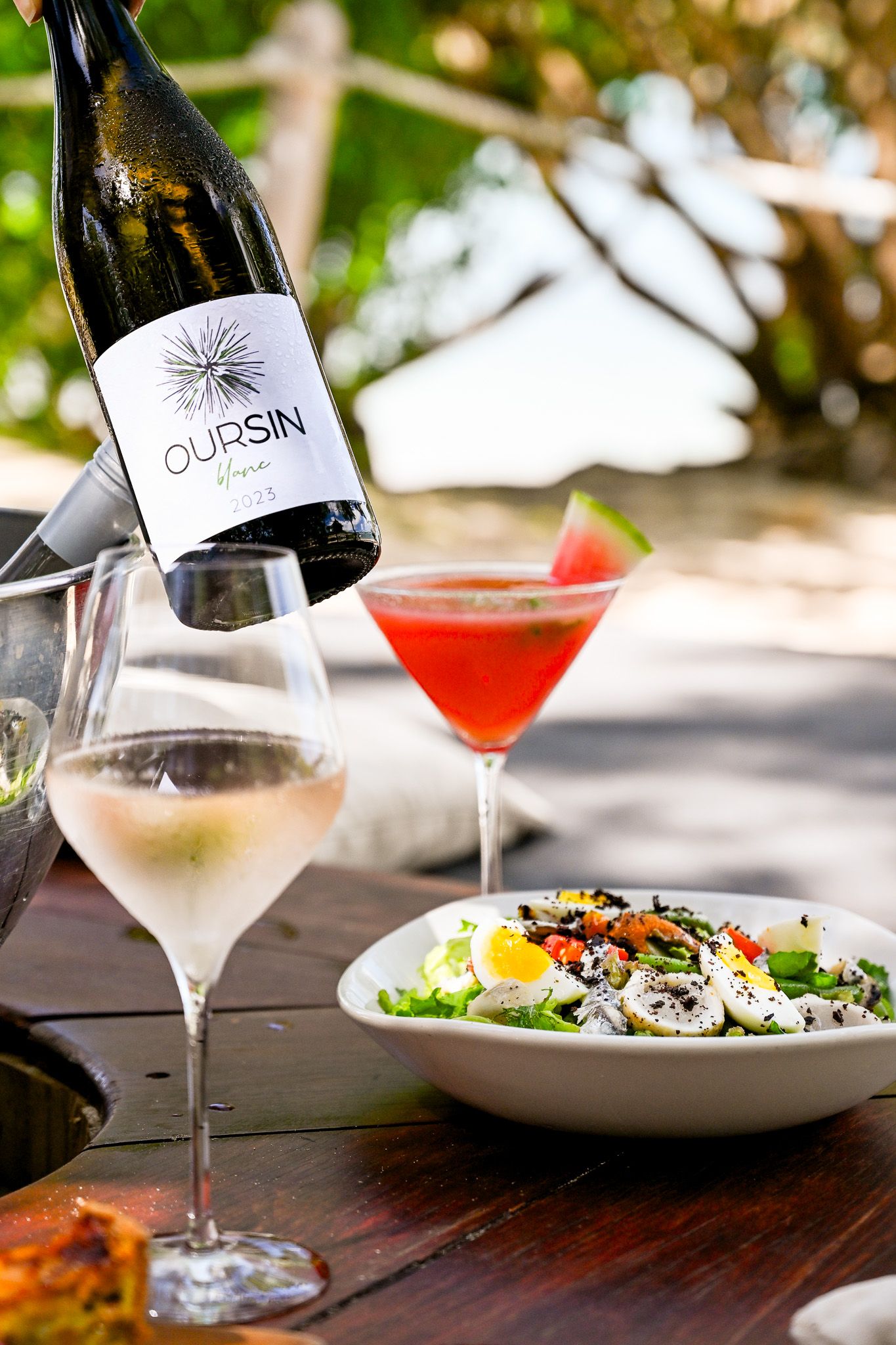Elegant cocktails and plated dishes arranged on a rustic wooden table with wine bottles in an outdoor dining setting.