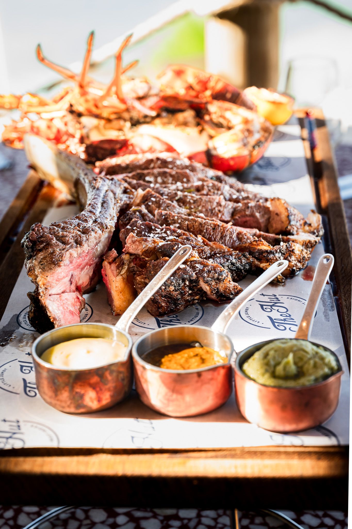 Grilled T-bone steak served with three copper pots of sauces and lobster tail on a rustic wooden board.