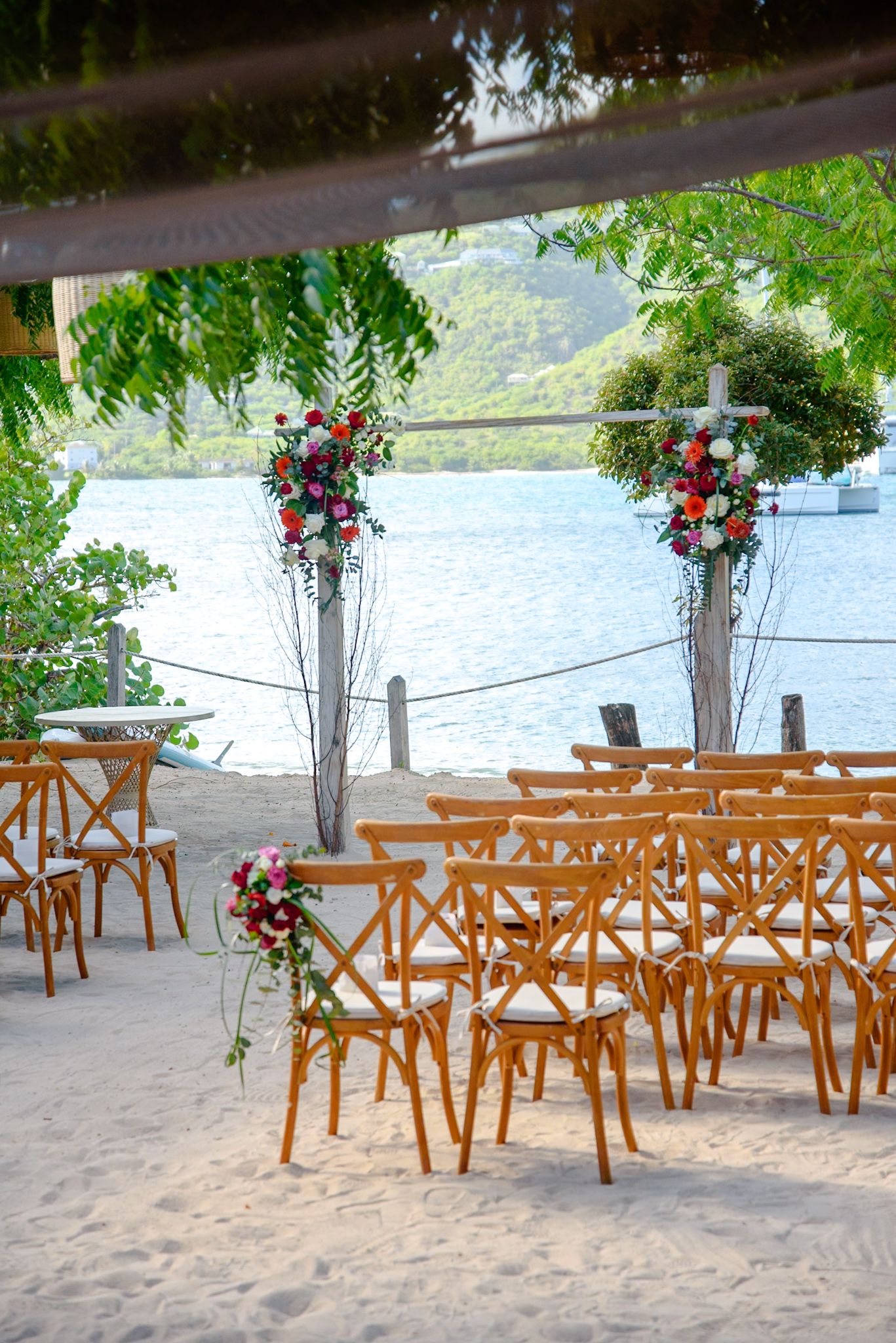 Wooden chairs arranged on a sandy beach for a waterfront wedding ceremony with tall floral arrangements in glass vases.
