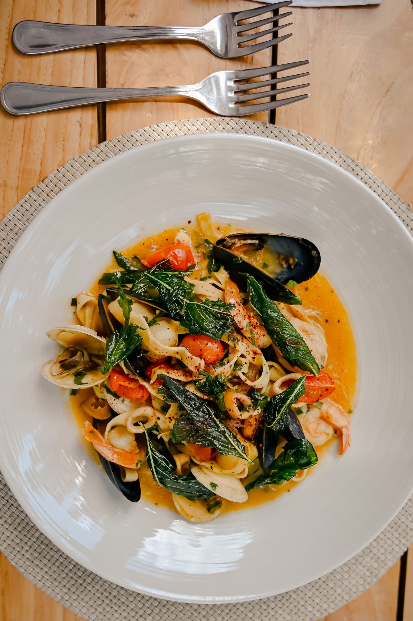 Three white plates of seafood pasta with shrimp, mussels and leafy greens in a light sauce arranged on a wooden surface.