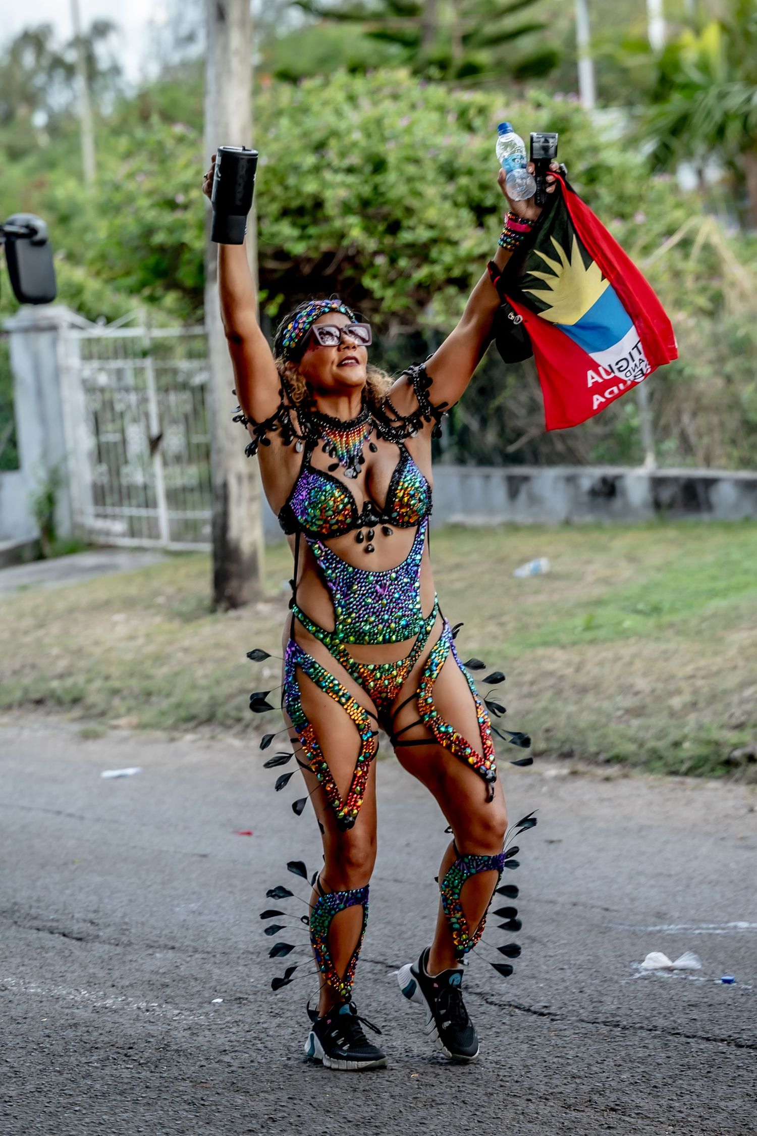 Woman in colorful carnival costume holds Antigua and Barbuda flag while dancing at outdoor festival celebration.