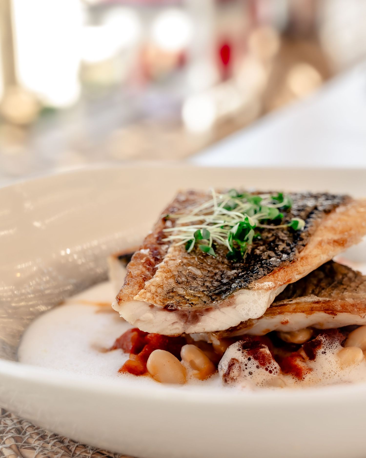 Pan-seared fish fillet with crispy skin, white beans, tomato sauce, and fresh microgreens on a white plate.