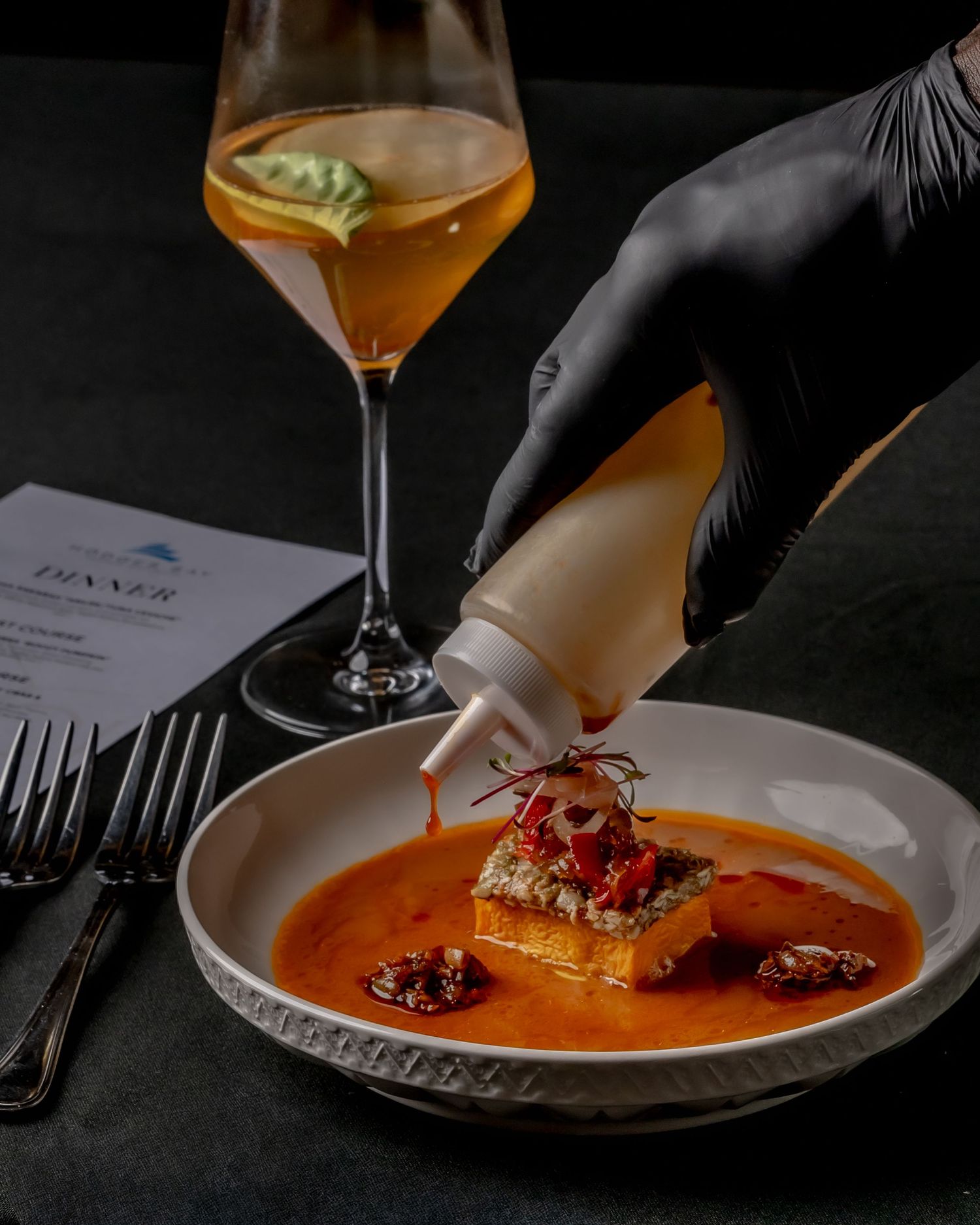 Chef in black gloves drizzling sauce on gourmet dish in white bowl, with orange cocktail in background.