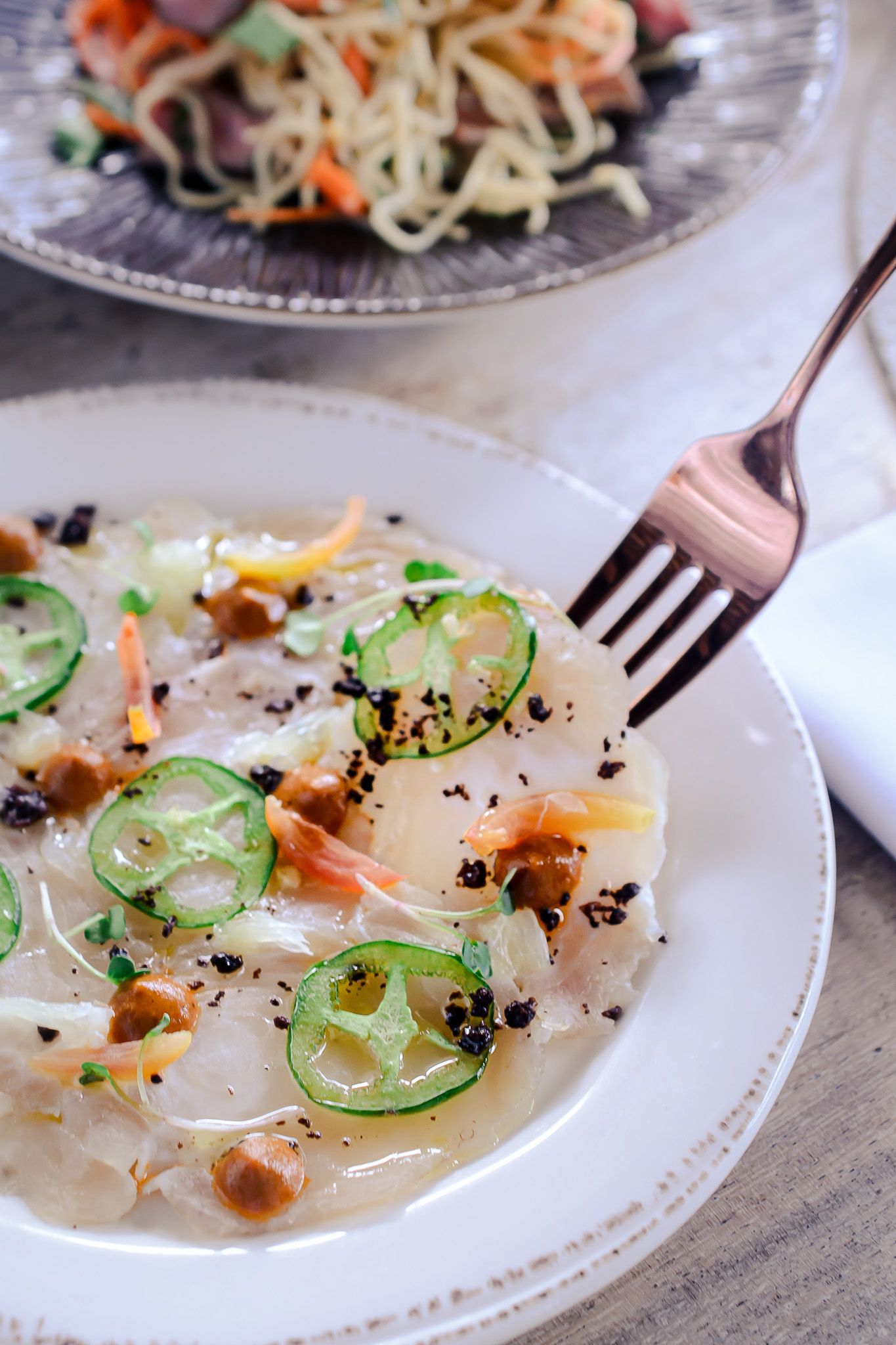 Thinly sliced raw fish crudo garnished with jalapenos, microgreens and black sesame seeds on white plates.