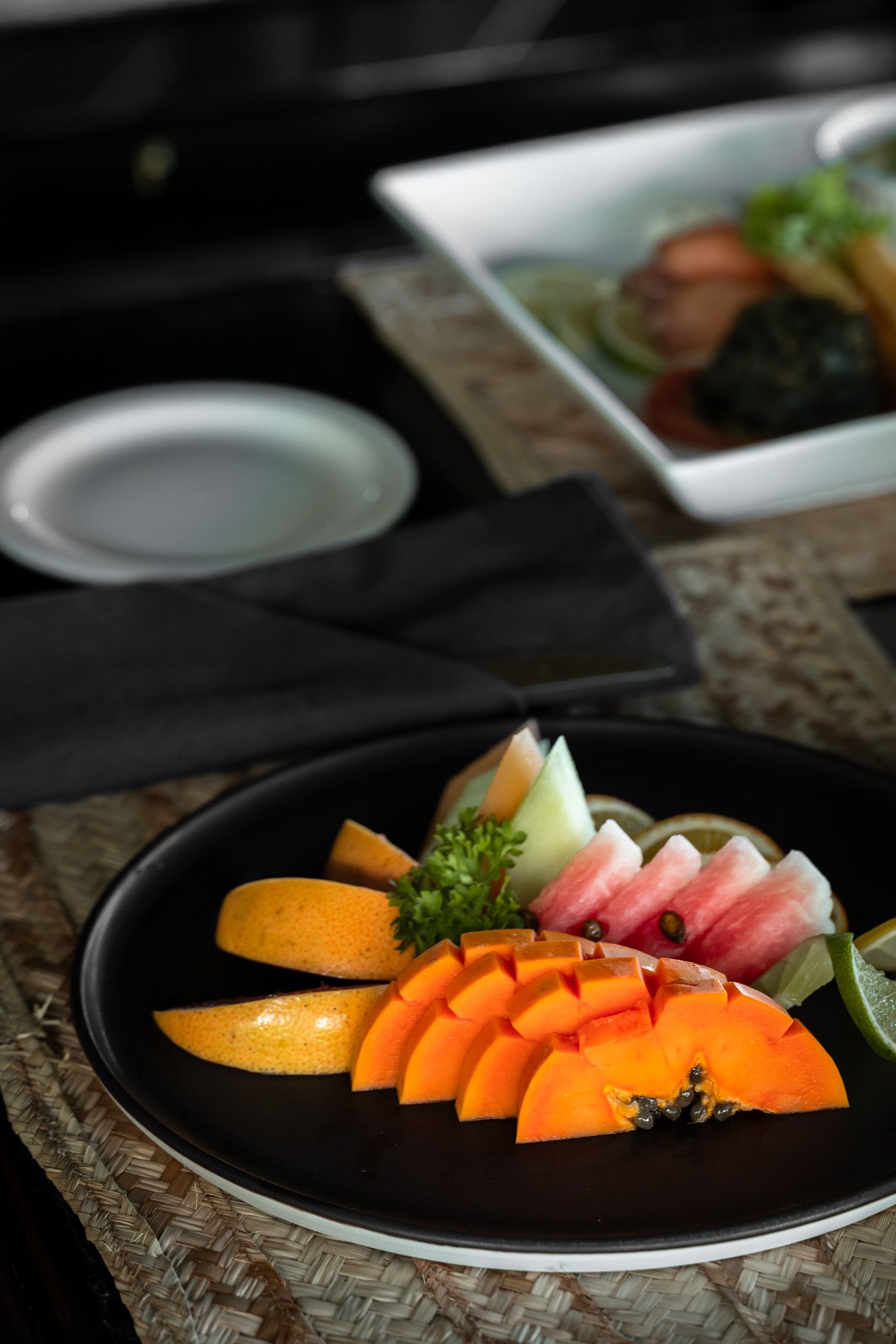 A black plate with sliced papaya, melon, and watermelon garnished with herbs, with a salad dish in the background.