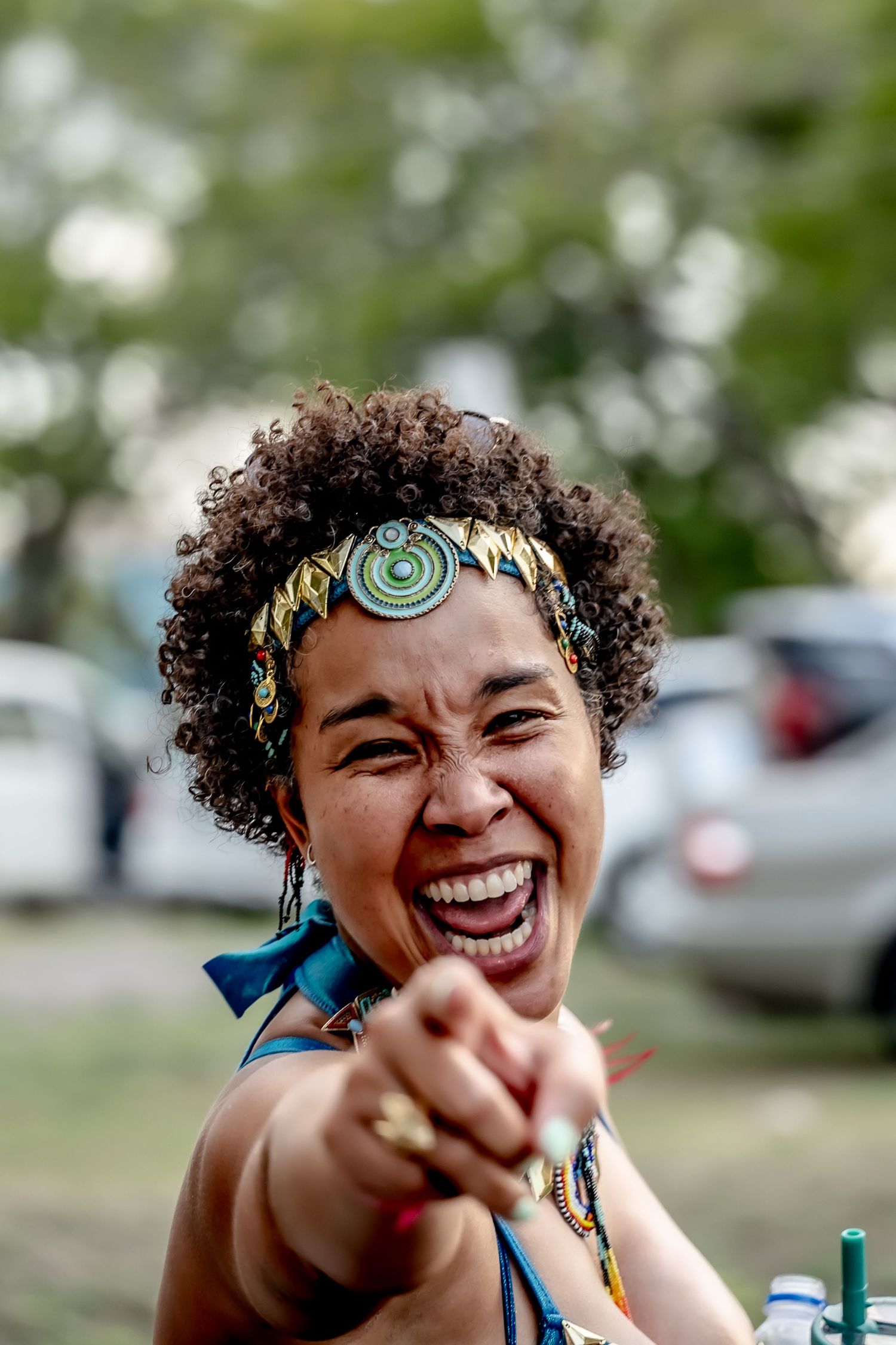 Joyful woman with curly hair and colorful headband laughing and pointing at camera outdoors.