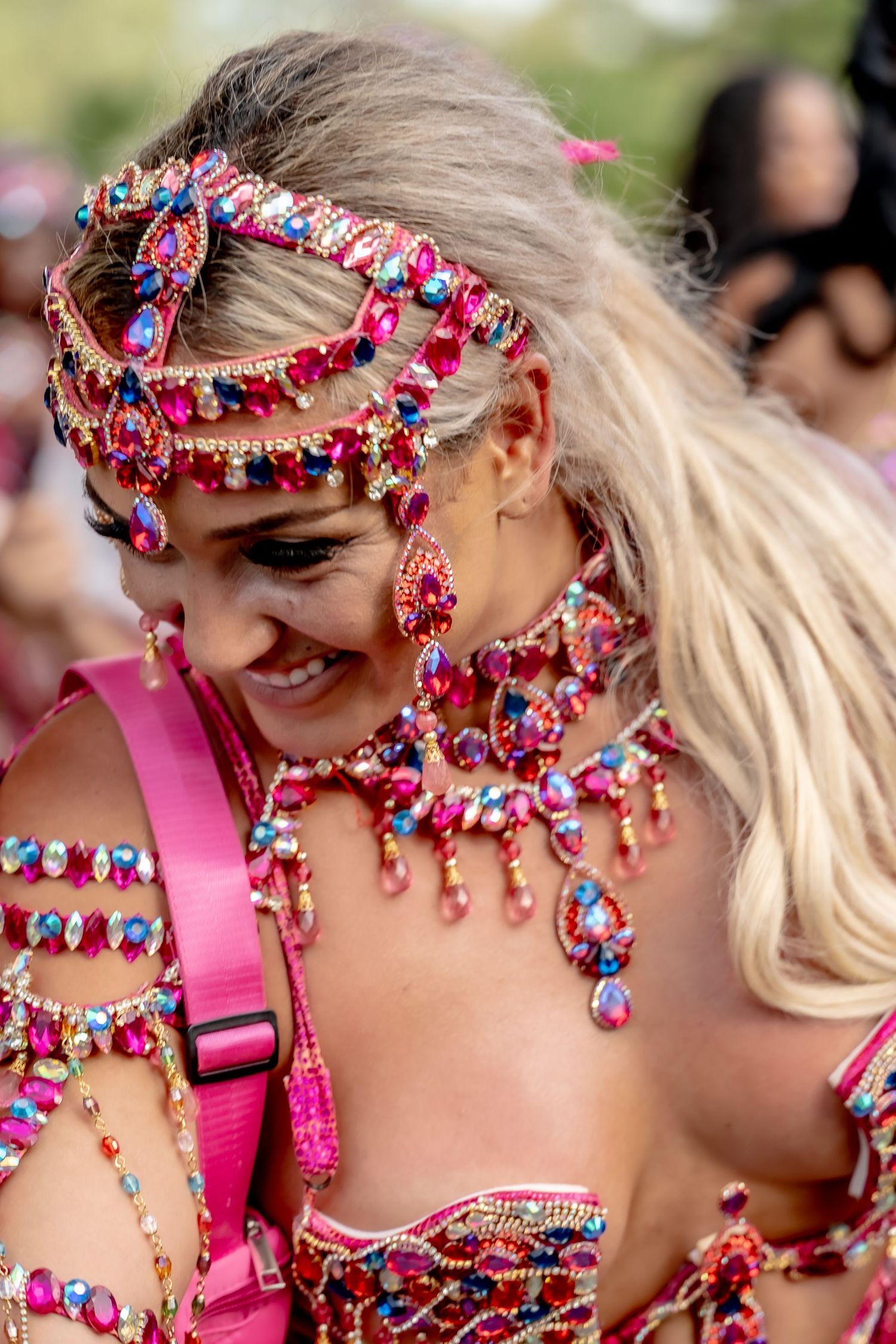 Woman in vibrant pink carnival costume adorned with colorful gemstone jewelry and headpiece, smiling at a festive event.
