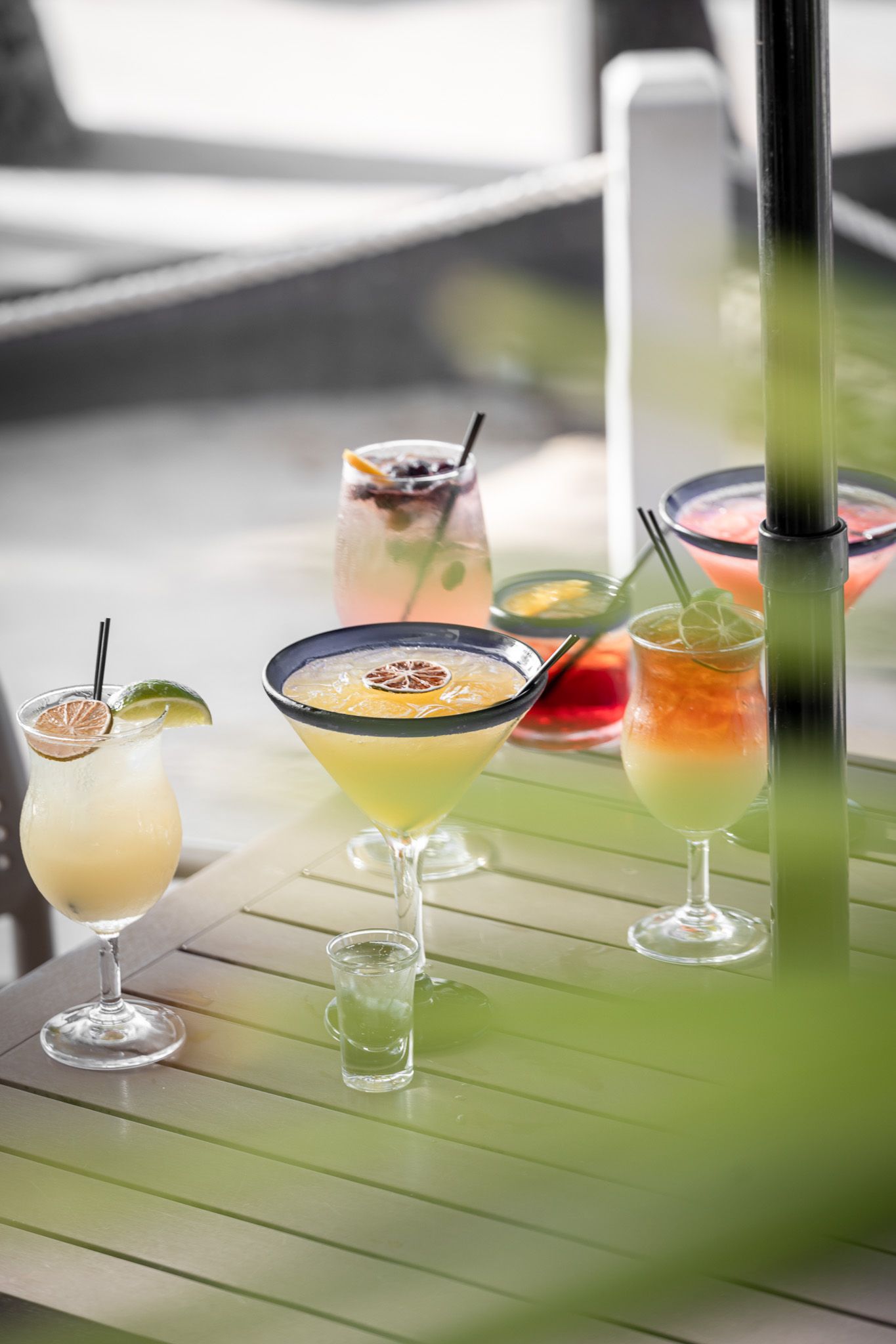 Colorful tropical cocktails with citrus garnishes arranged on a green outdoor table at a sunny bar.