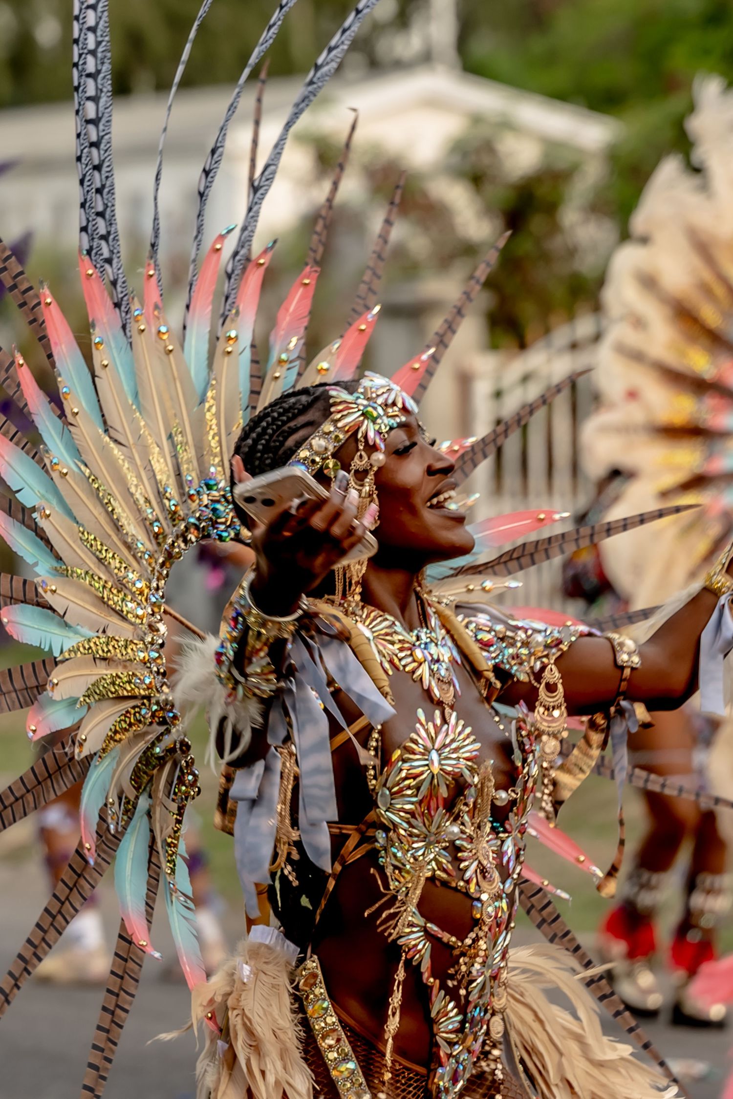 A woman in a vibrant feathered carnival costume with gold embellishments dances joyfully during a colorful street parade.