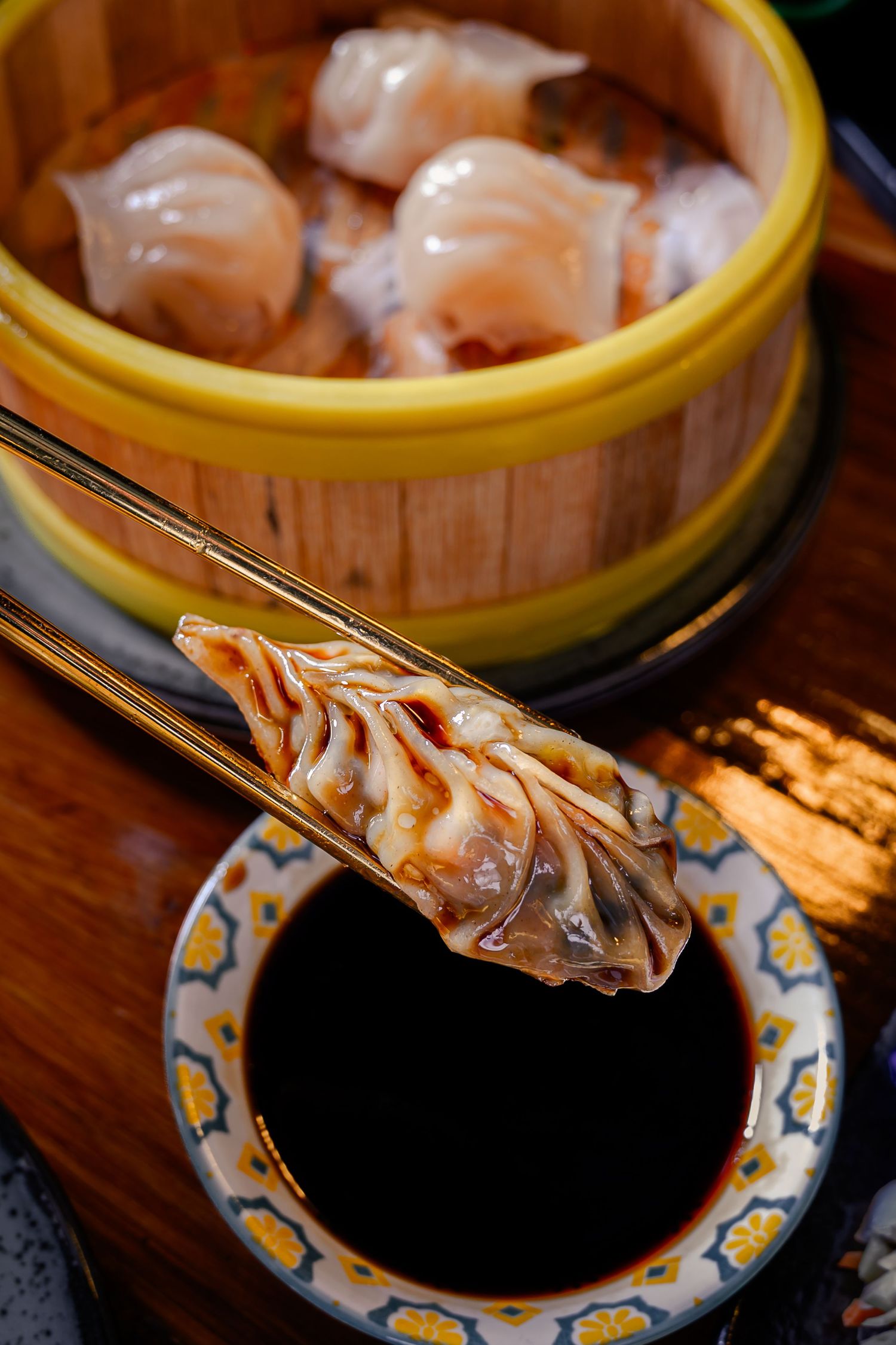 Chopsticks holding a shrimp dumpling over soy sauce with a bamboo steamer basket of dim sum in the background.