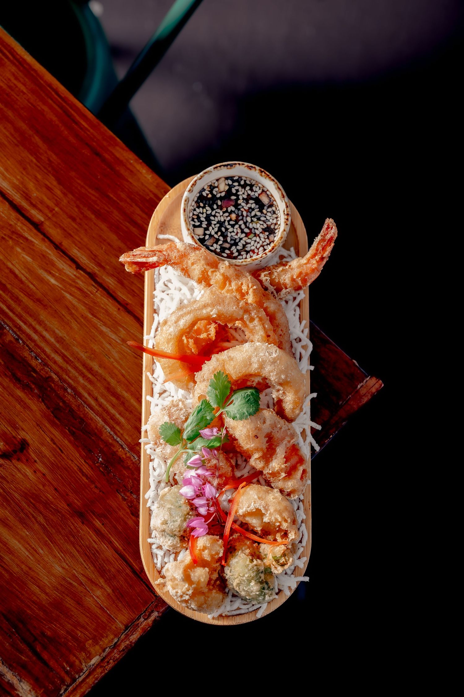 Tempura shrimp served in an elongated wooden boat with dipping sauce topped with sesame seeds on a rustic table.