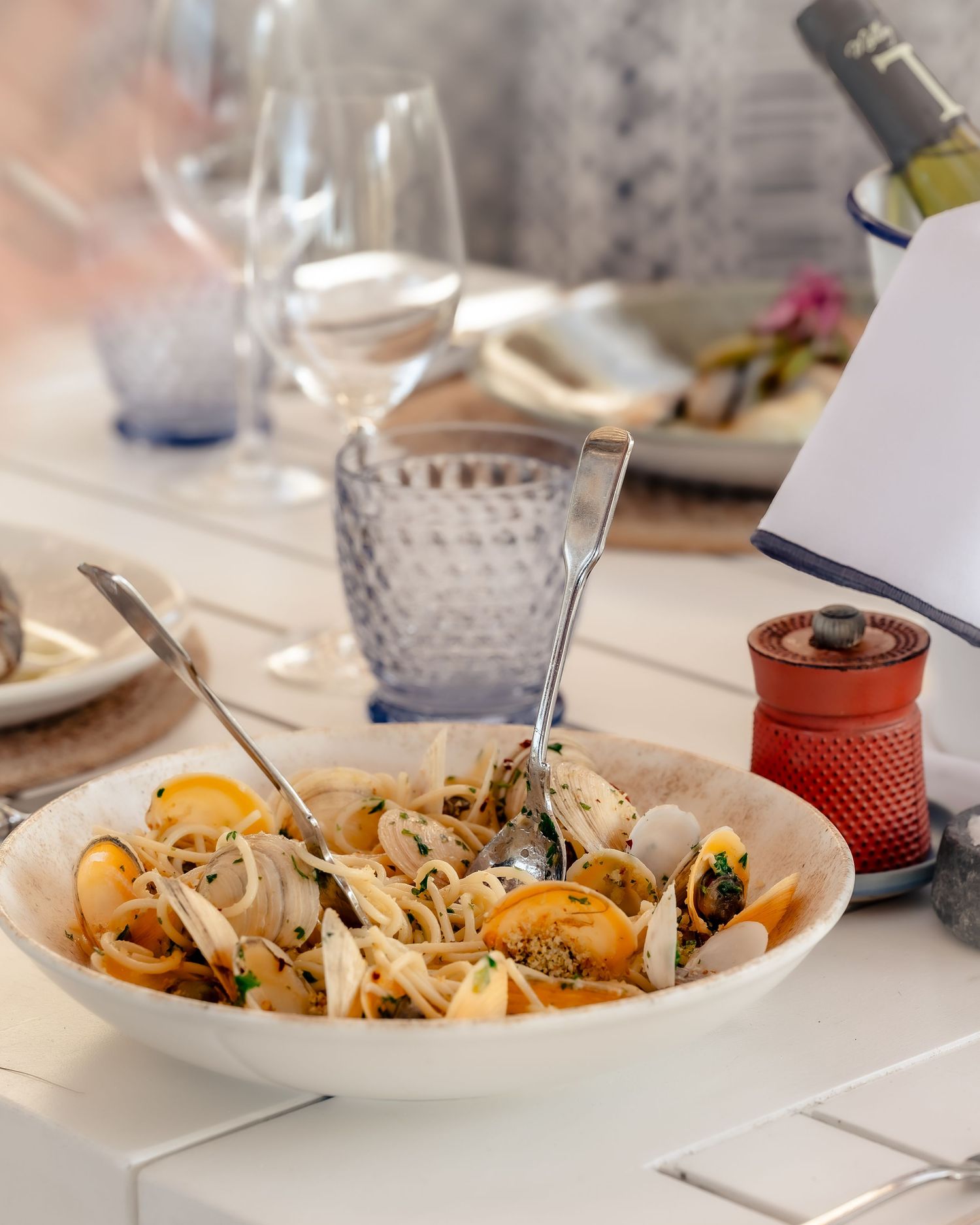 A white bowl of clams with pasta and two spoons on a restaurant table with wine glasses.
