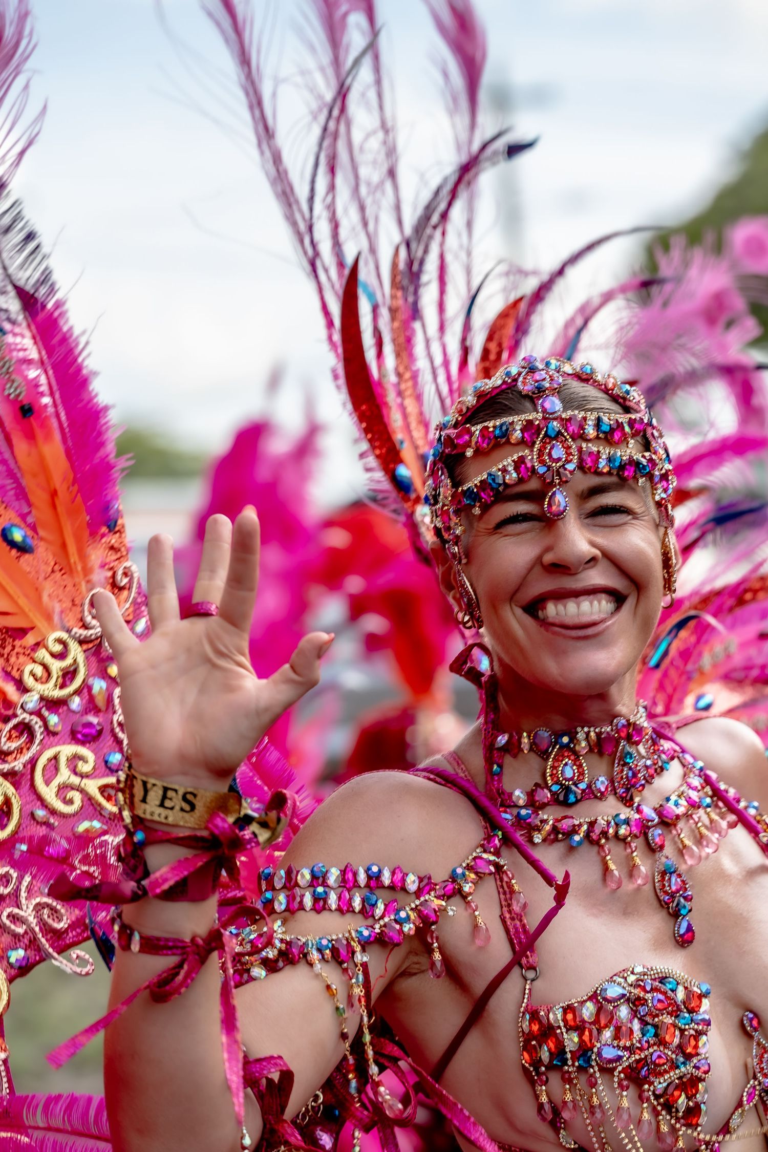 Smiling woman in vibrant pink feathered carnival costume with jeweled headdress and beaded bra waving at parade.